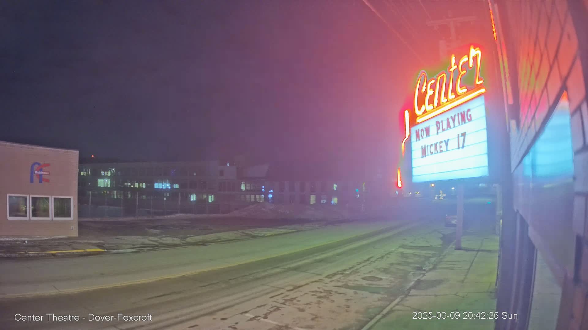 A foggy night scene shows a street with a movie theater marquee displaying "Mickey 17,"  and buildings in the background.