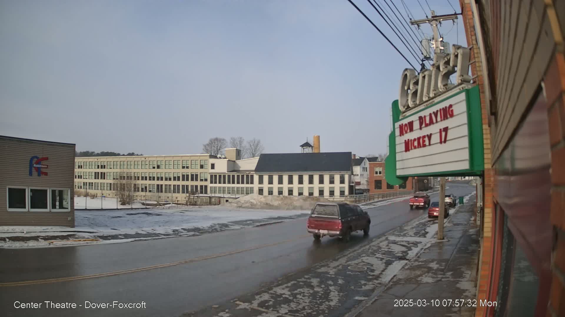 A partly sunny day reveals a street scene with melting snow and ice on the road, several buildings including a large multi-story structure and a smaller brick building with a marquee, and a few vehicles driving by.