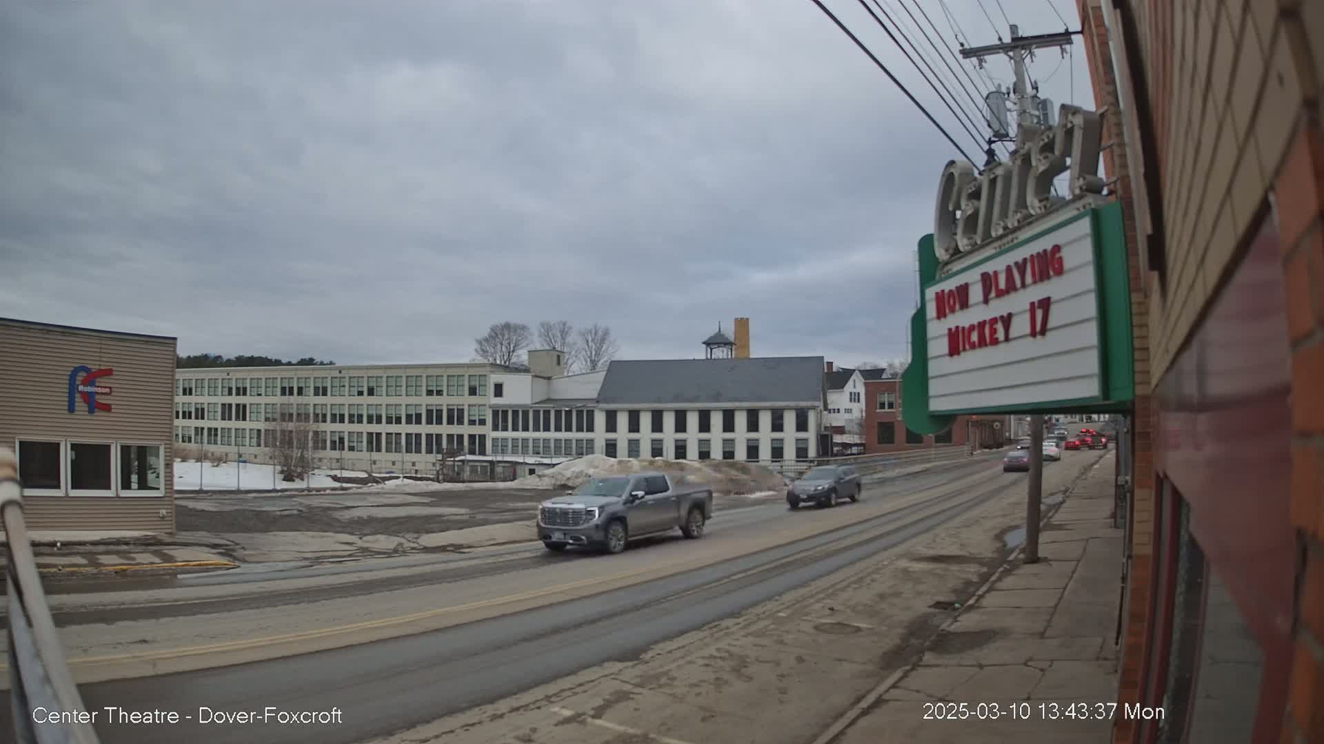 A street scene on an overcast day shows a silver pickup truck and other vehicles driving past a large, multi-story building and a movie theater marquee.