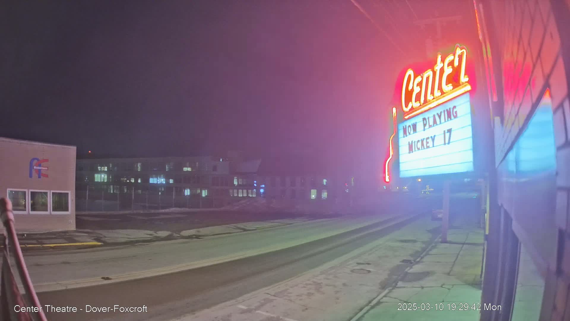 A foggy night scene shows a movie theater marquee advertising "Mickey 17," with buildings and a street visible in the background.