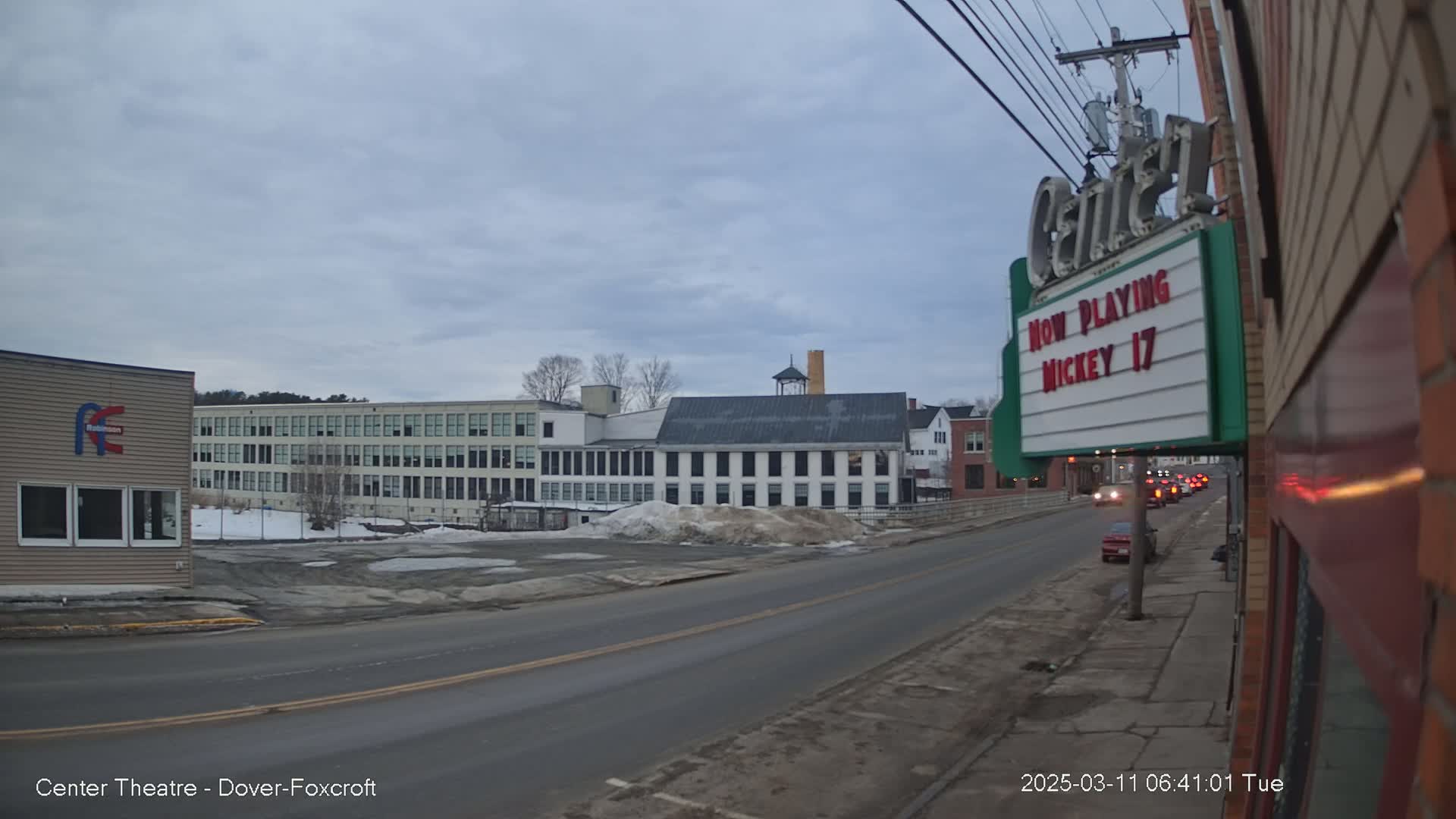 A street scene under a cloudy sky shows several buildings, including a large multi-story factory-like structure, a smaller building with a marquee that says "Now Playing Mickey 17", and a few cars driving down the street.