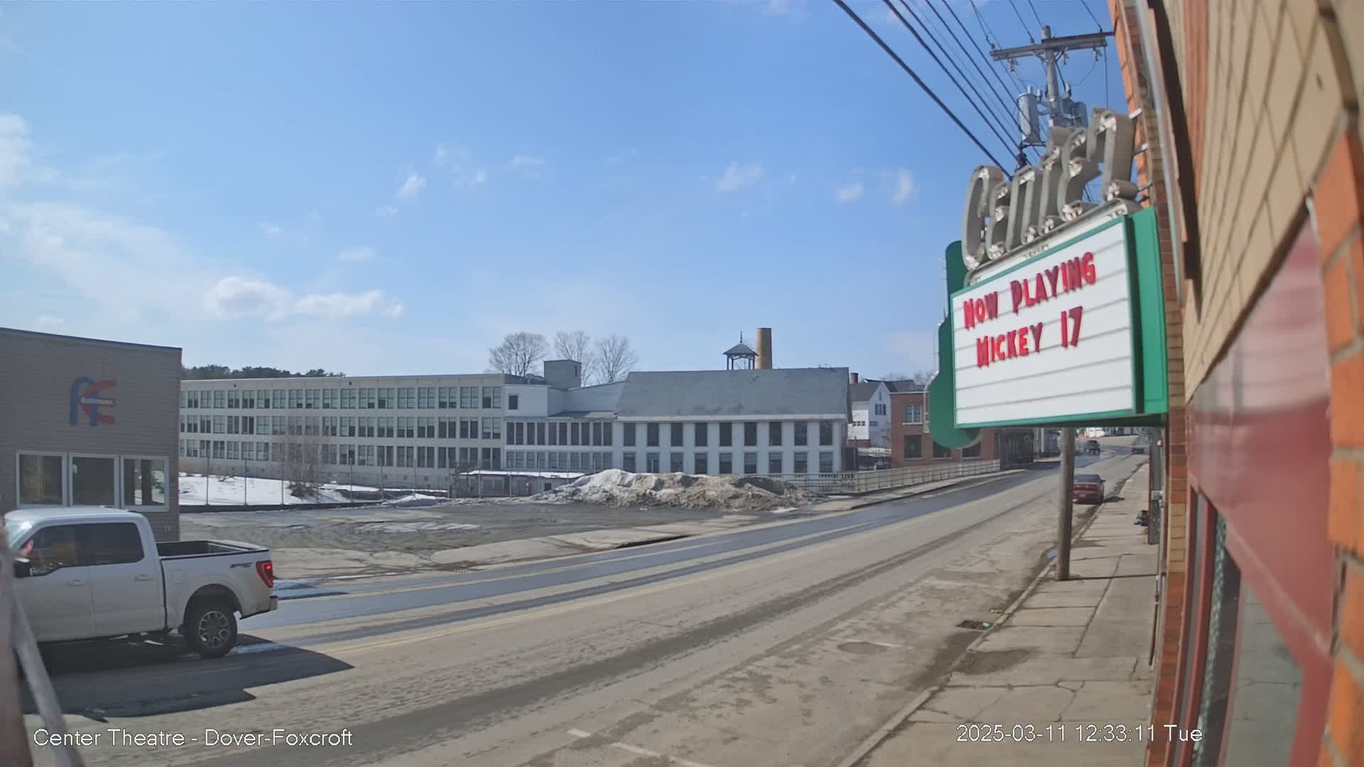 A sunny day reveals a street scene with a large building and a movie theater marquee showing "Mickey 17," and a pickup truck parked on the side of the road.
