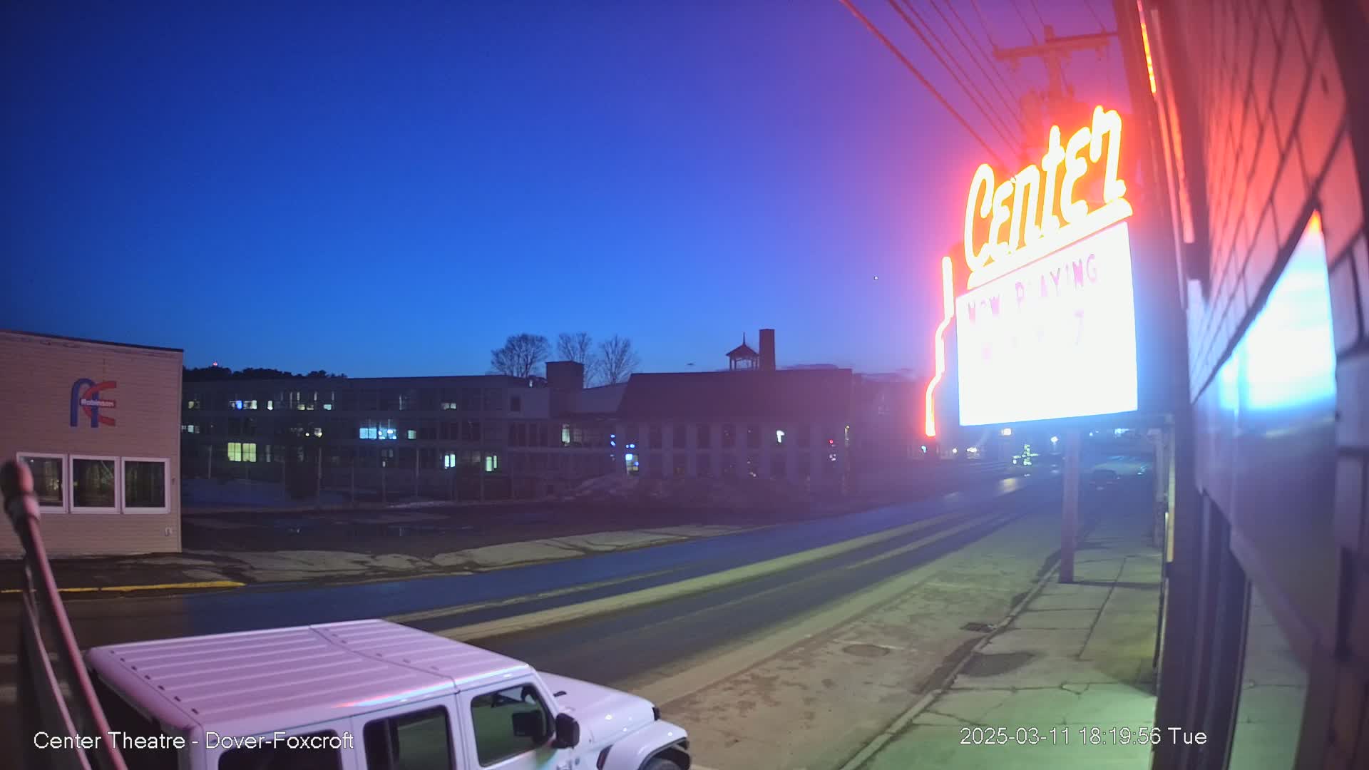 A nighttime street scene shows a road leading towards a few buildings under a twilight sky, with a neon sign for a theater visible on the right.