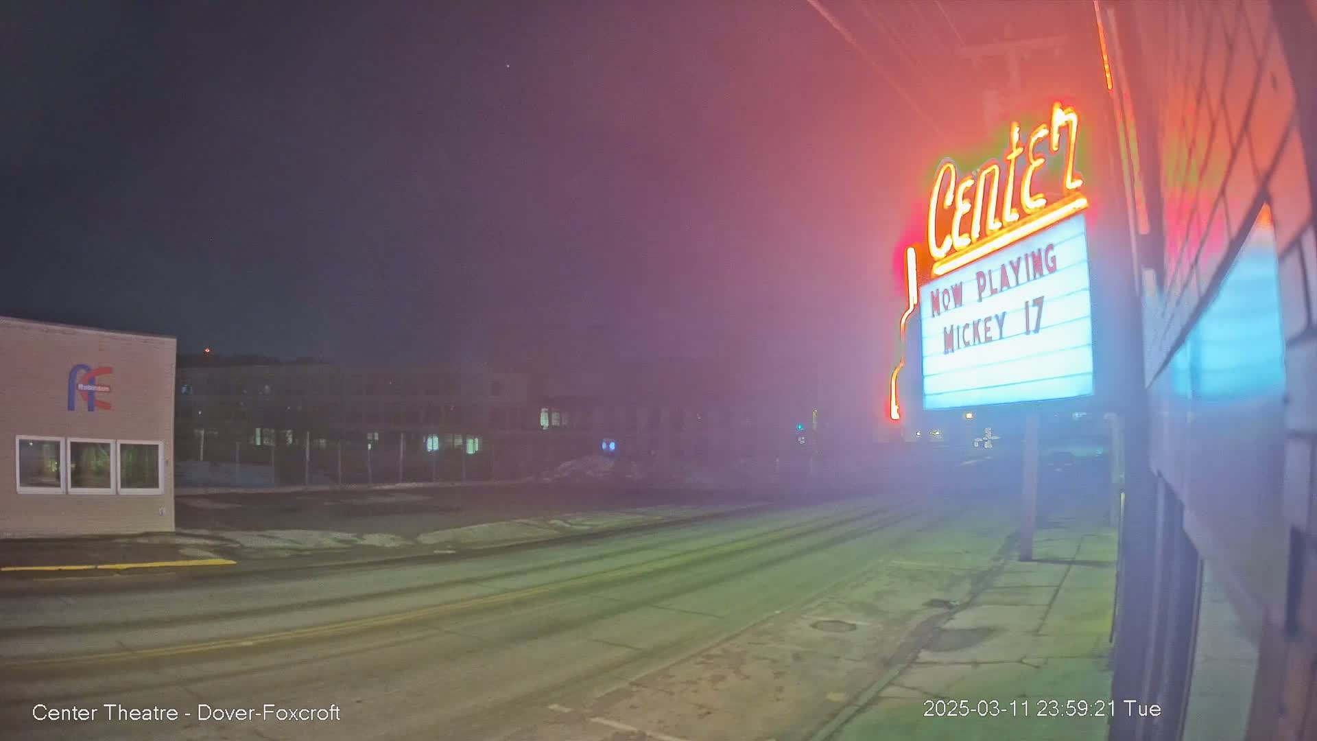 A foggy, nighttime street scene shows a movie theater marquee advertising "Mickey 17," with buildings and a road visible in the background.