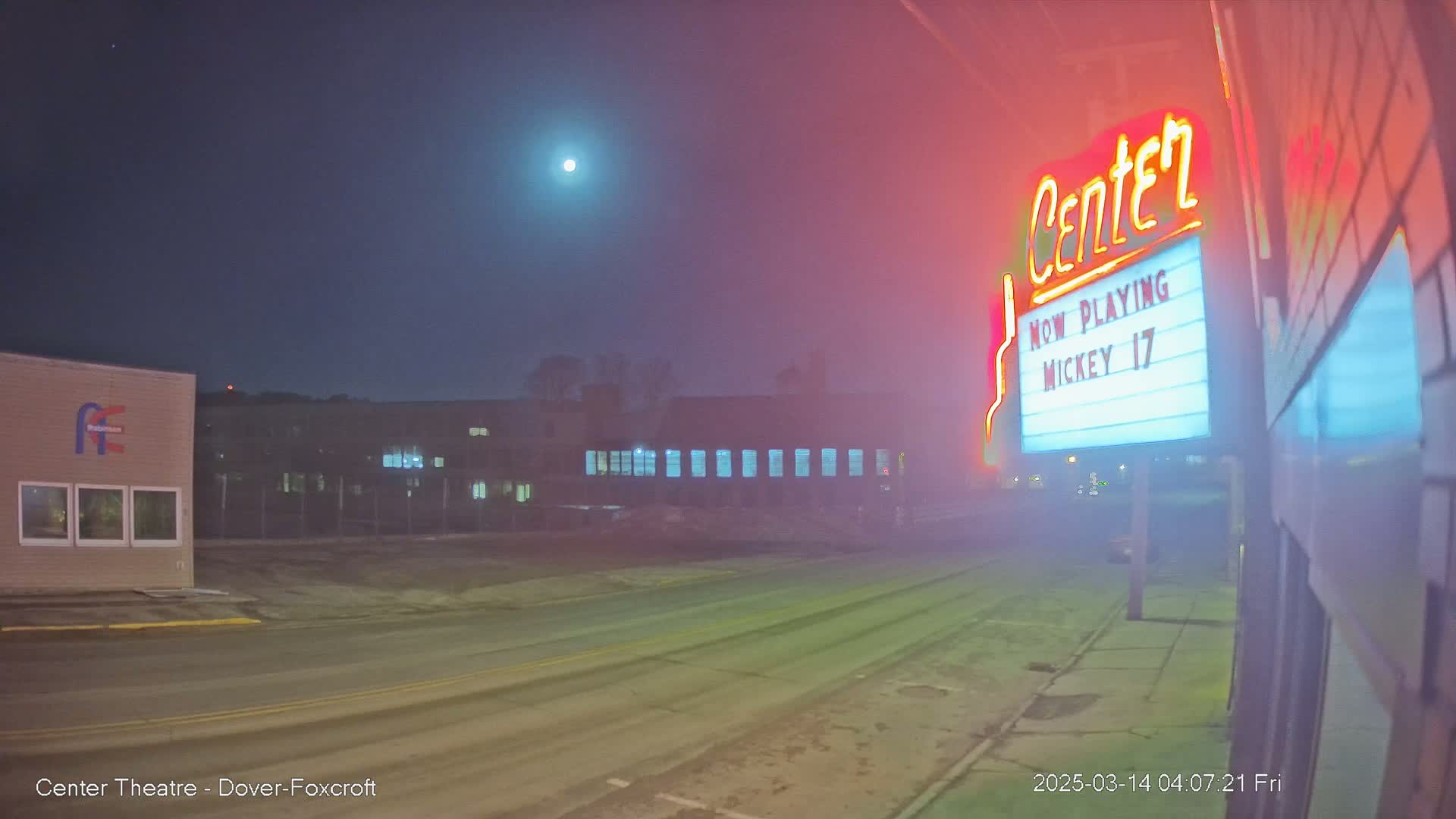 A foggy night scene shows a street with a building and a movie theater marquee advertising "Mickey 17," under a bright moon.