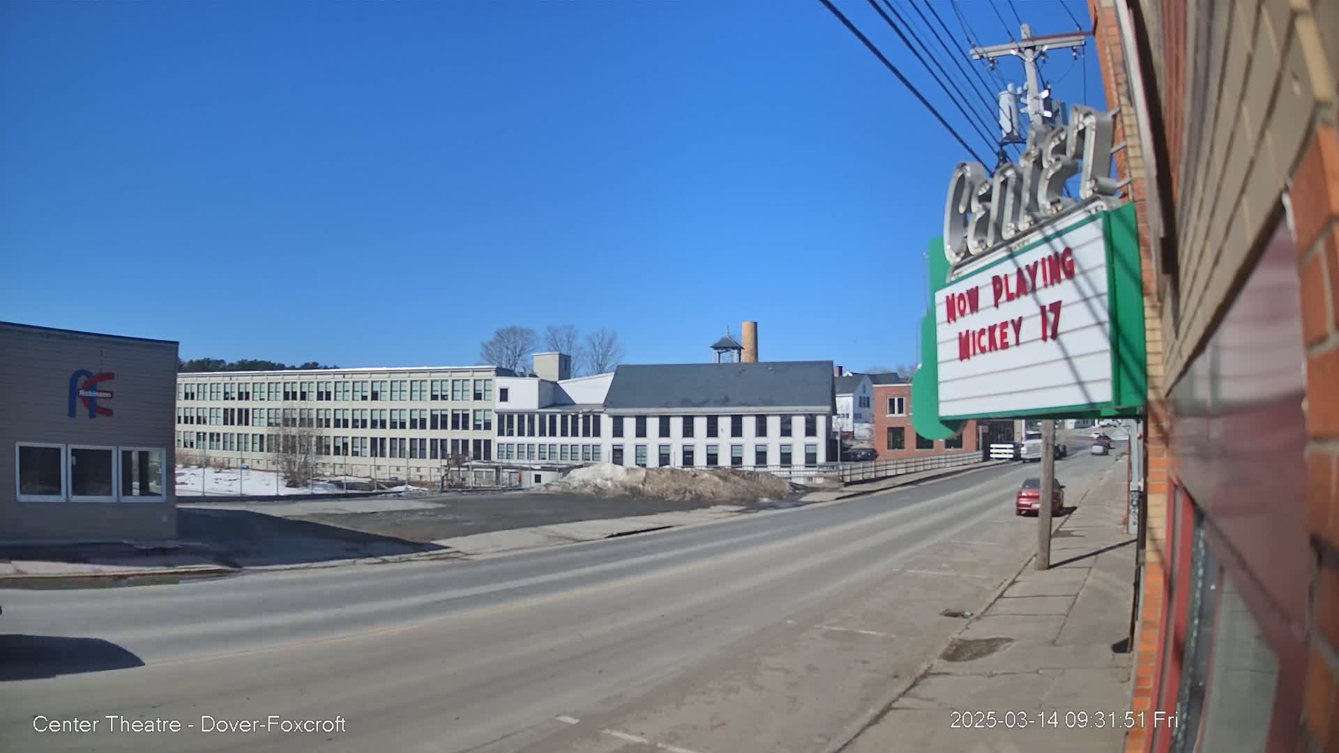 A sunny day reveals a street scene with several industrial-style buildings, a small empty lot, and a few cars parked along the roadside.