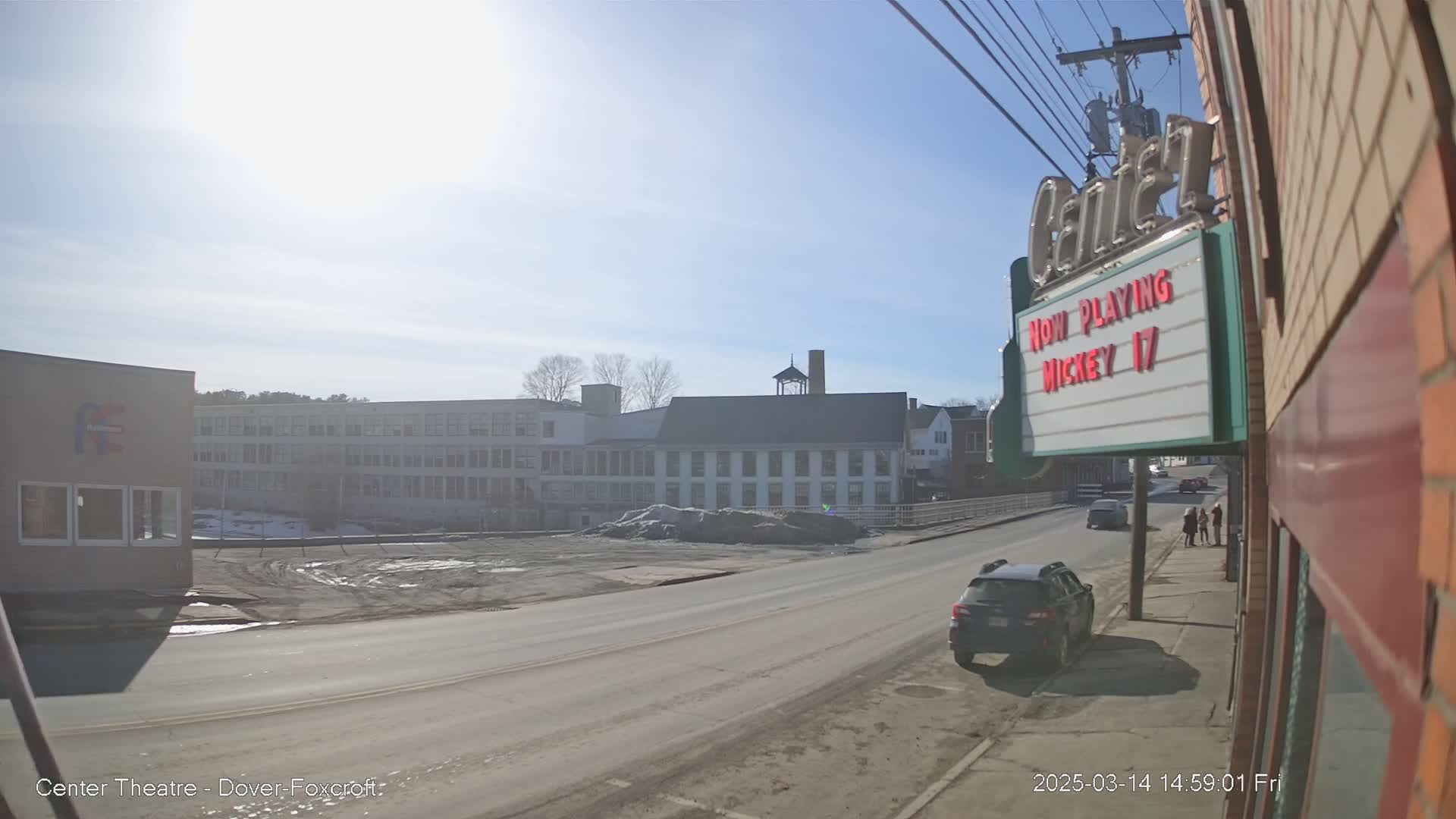 A sunny day reveals a street scene with a multi-story factory building, a smaller building with a marquee, and a few cars parked along the road.
