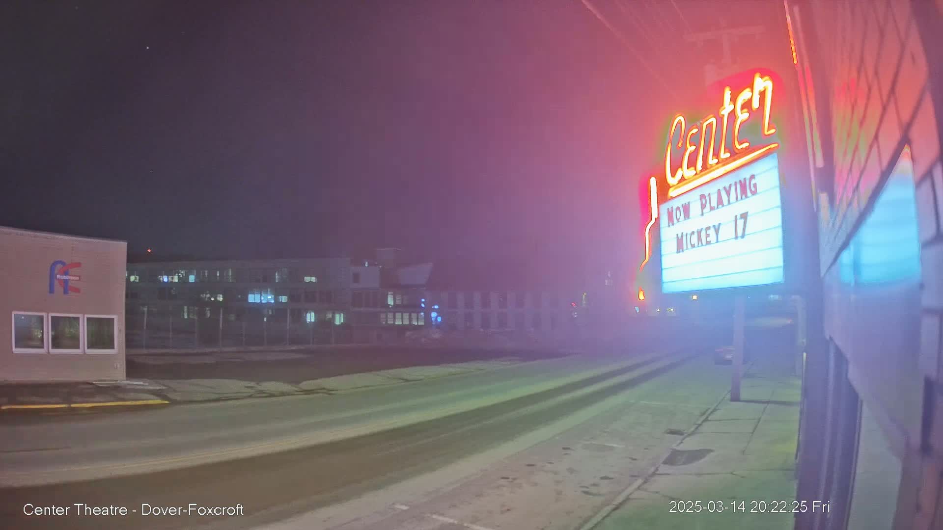 A snowy street at night, illuminated by the neon lights of a movie theater marquee advertising "Mickey 17".