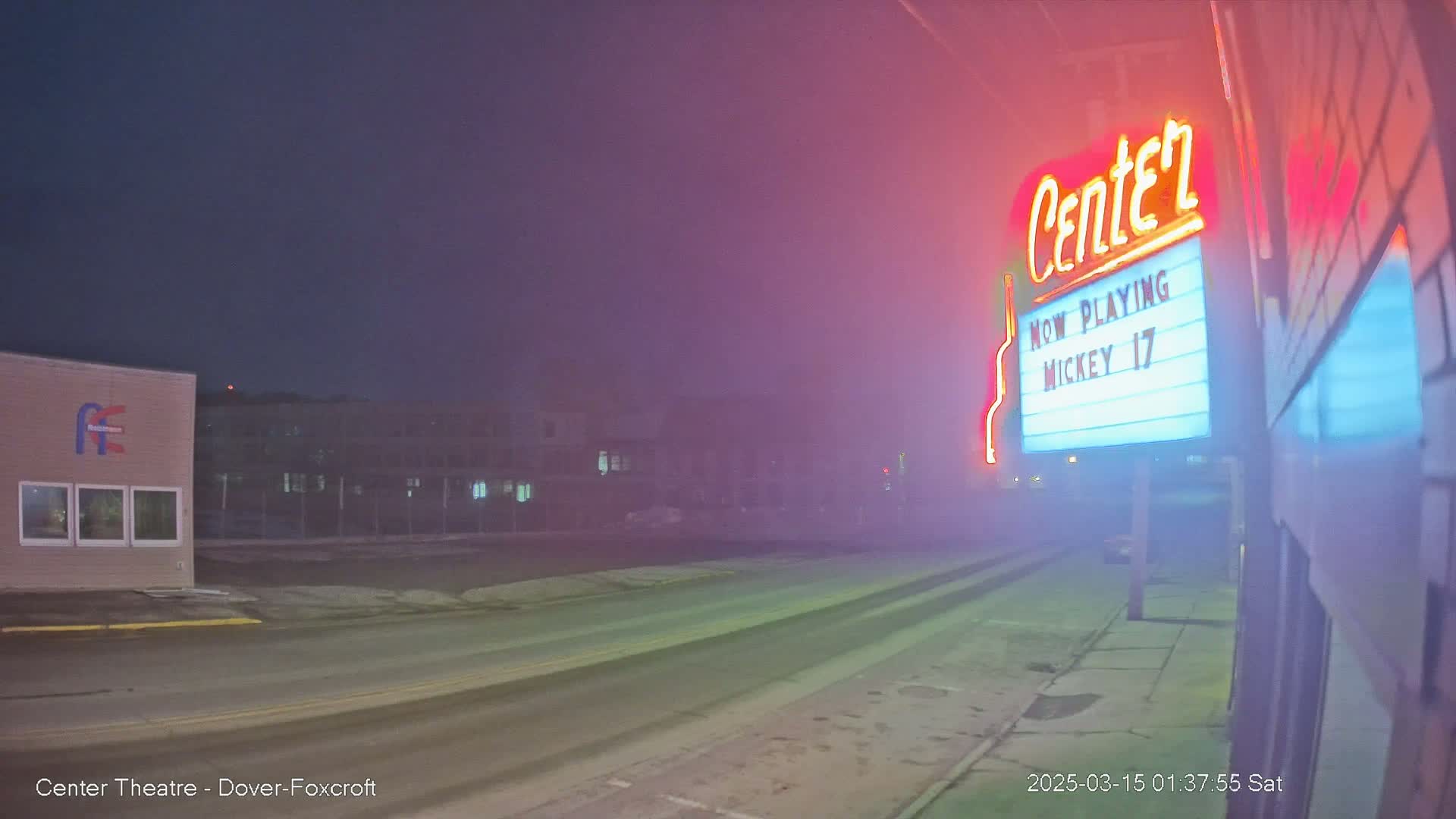 A foggy, nighttime street scene shows a movie theater marquee advertising "Mickey 17," with a building and some cars visible in the distance.