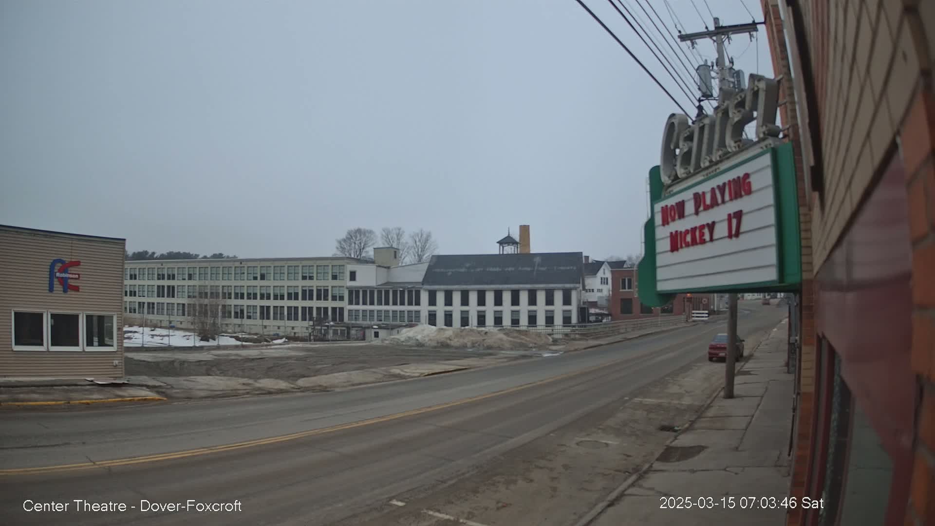A mostly empty street on an overcast day leads to several large buildings, one of which is a movie theater.