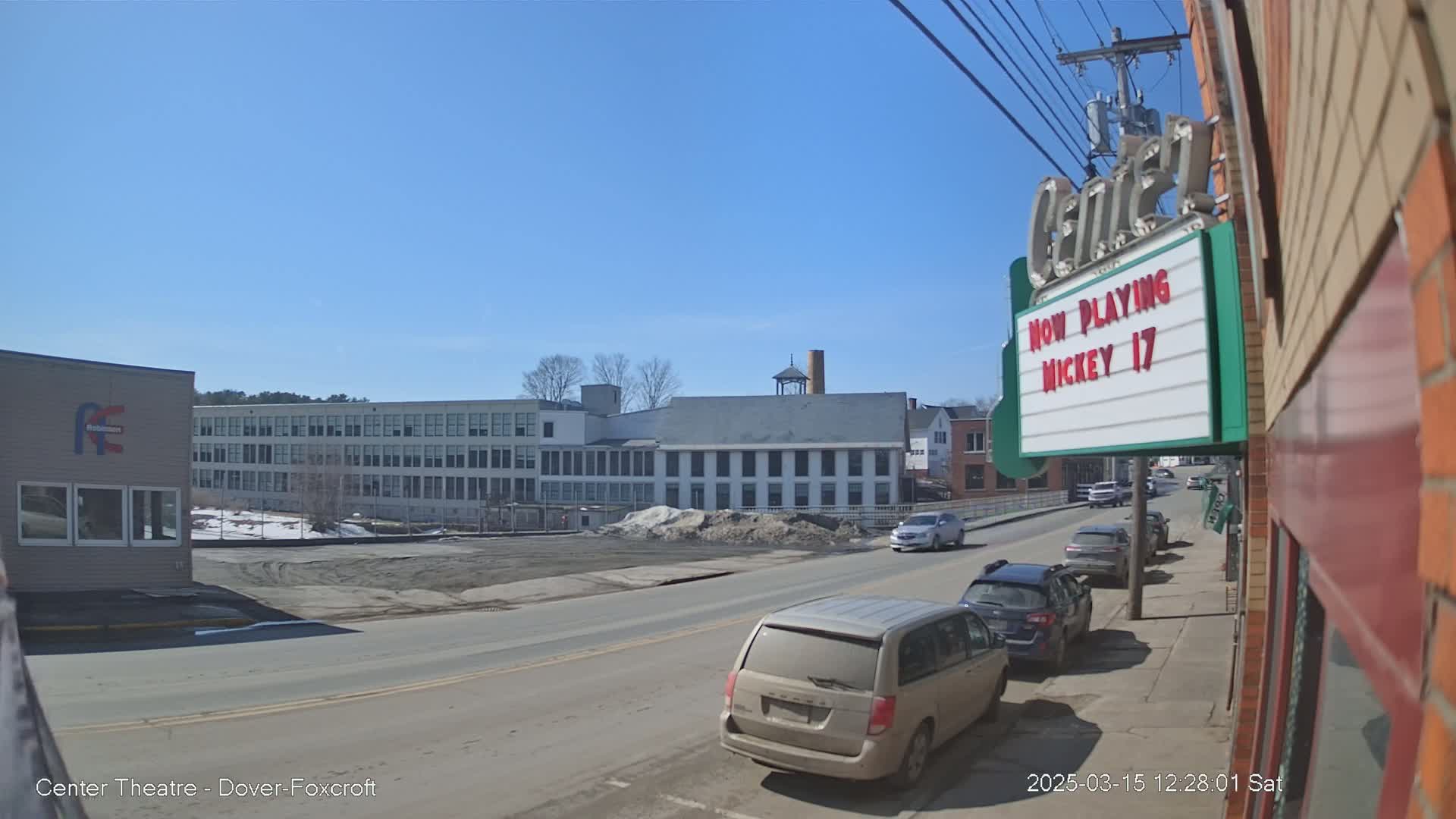 A street scene on a sunny day shows several cars parked along a street next to a brick building with a marquee that says "Now Playing Mickey 17," and across the street are several multi-story buildings.