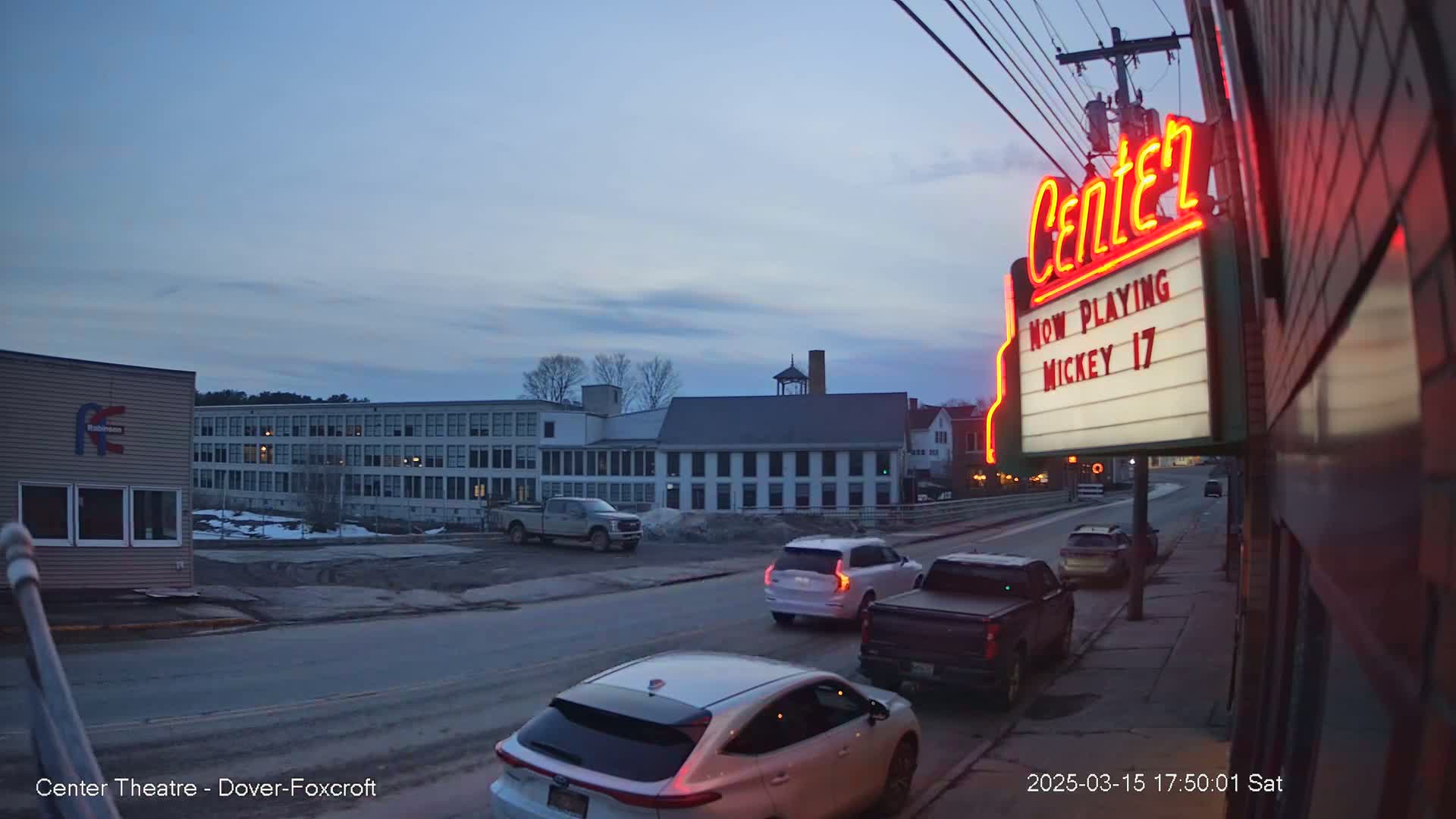 A street scene at dusk shows several vehicles parked and driving near a movie theater marquee with a lit sign.