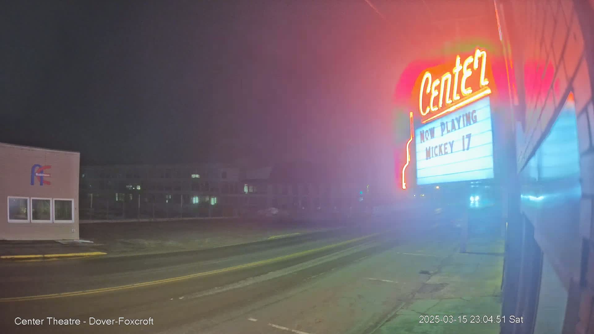 A foggy night scene shows a movie theater marquee with a neon sign, and a street with buildings in the background.