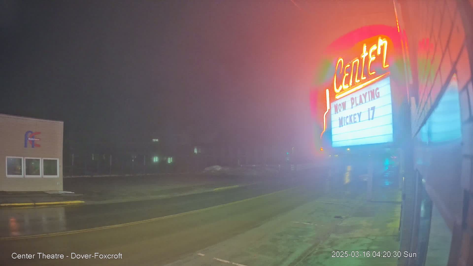 A fog-shrouded street at night shows a movie theater marquee advertising "Mickey 17," with a building across the street visible in the background.