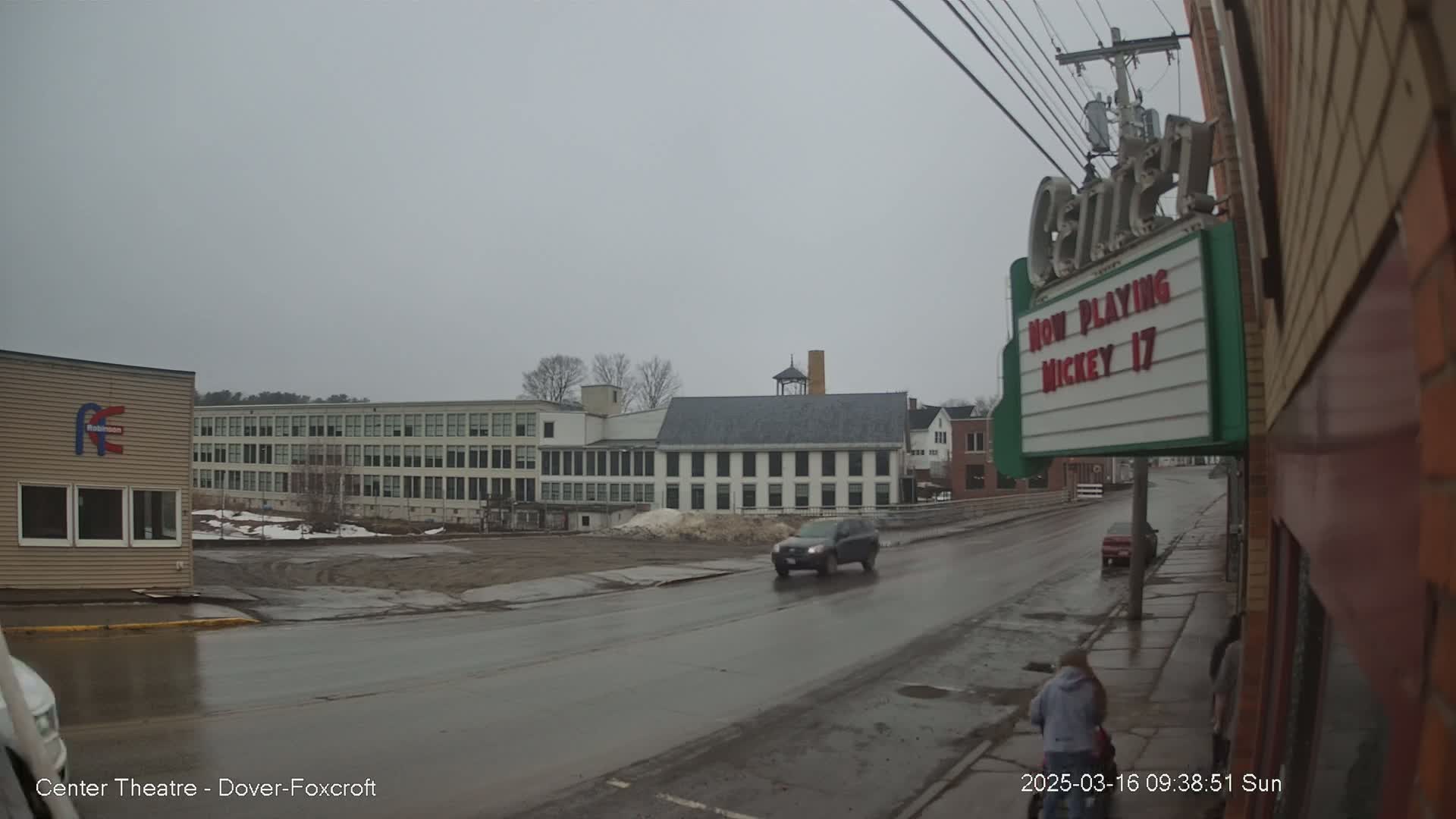 A wet street scene on an overcast day shows a dark-colored SUV driving past buildings, including a multi-story factory and a movie theater marquee.