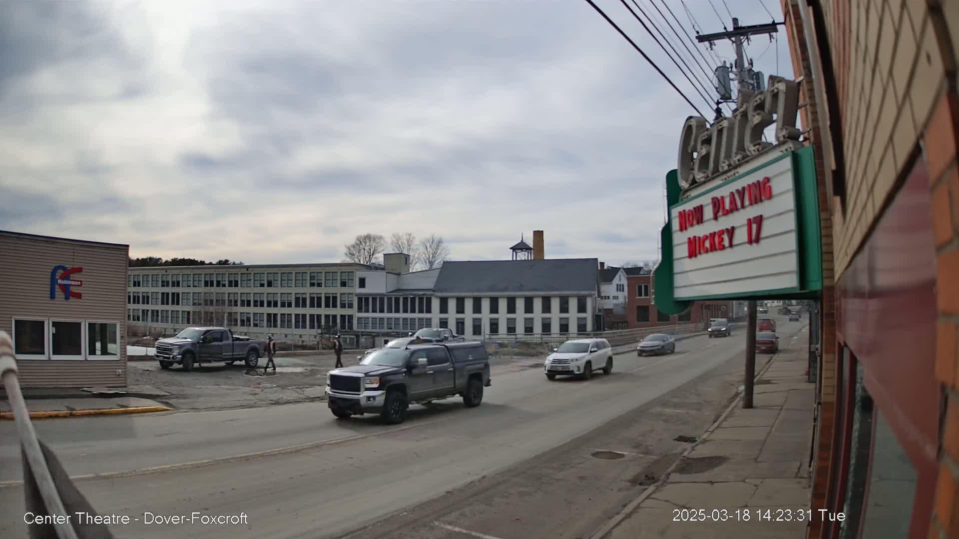 A street scene on an overcast day shows several vehicles driving past a large industrial building and a movie theater marquee.