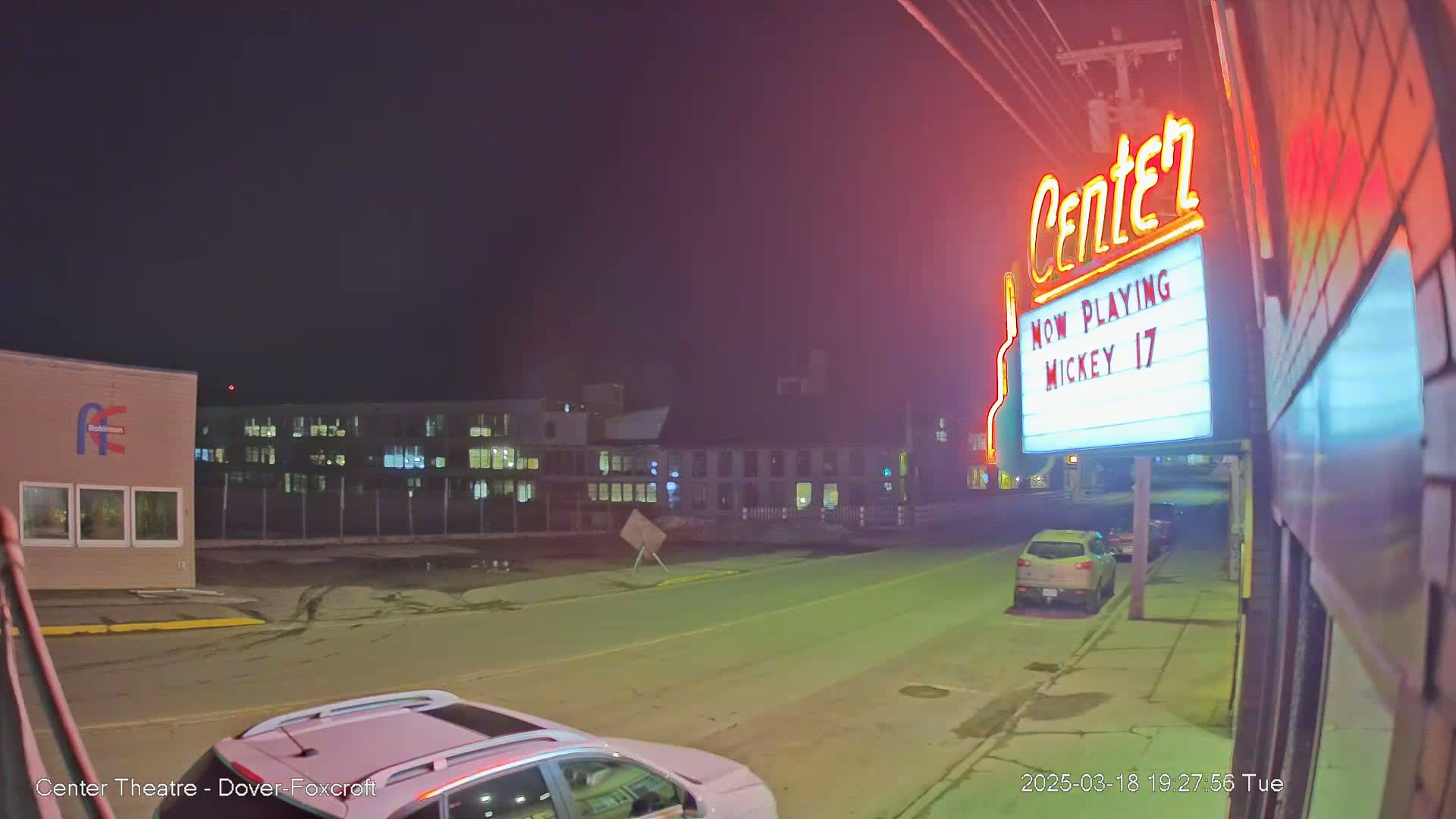 A nighttime street scene shows a movie theater marquee advertising "Mickey 17," with parked cars and buildings visible under a dark sky.