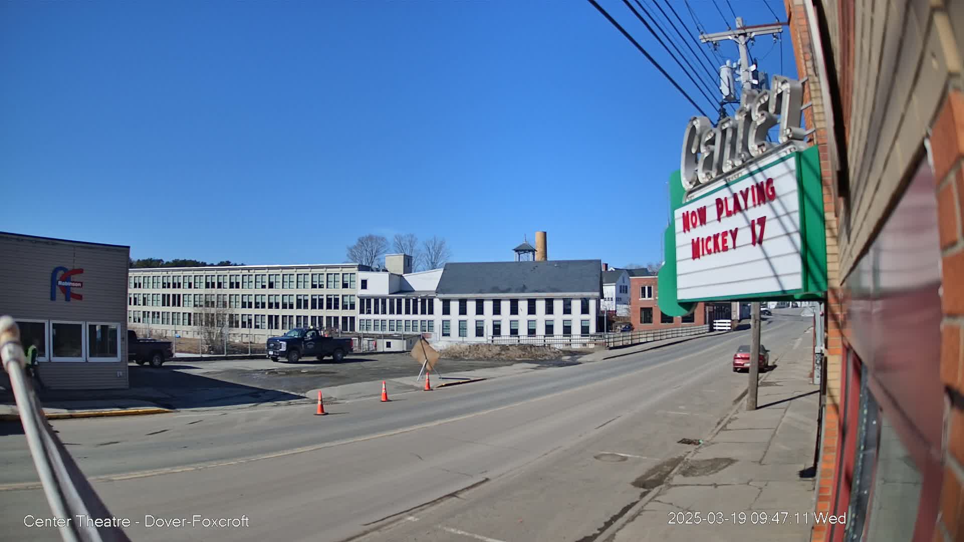 A street scene on a sunny day shows a row of industrial buildings, a few vehicles, and a movie theater marquee.