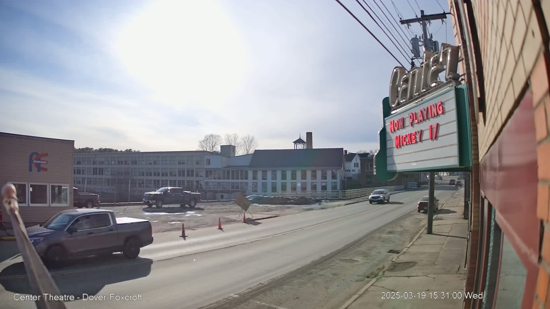 A sunny day reveals a street scene with several vehicles, including pickup trucks, and buildings including a multi-story factory and a movie theater showing "Mickey 17."