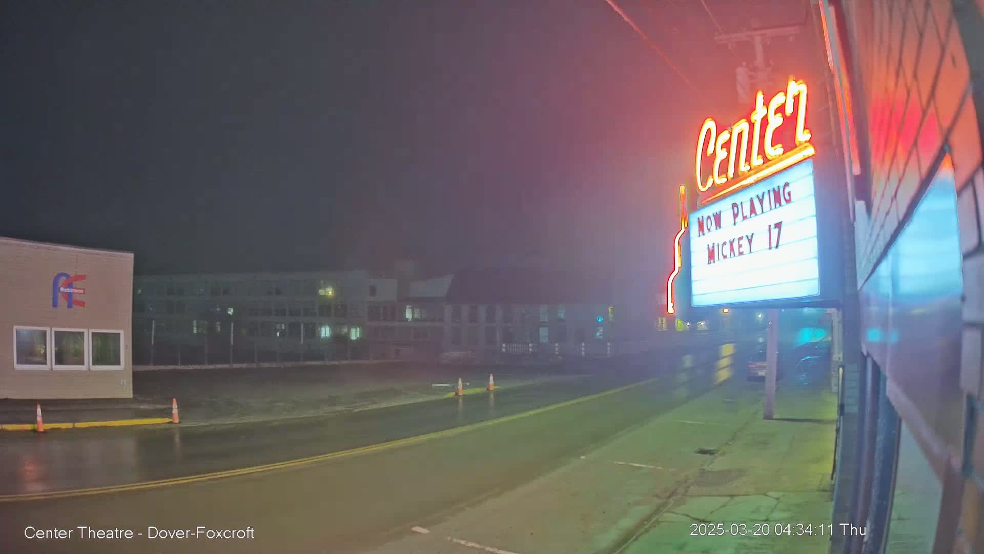 A rain-slicked street at night, illuminated by the neon sign of a movie theater showing "Mickey 17," with a building in the background.
