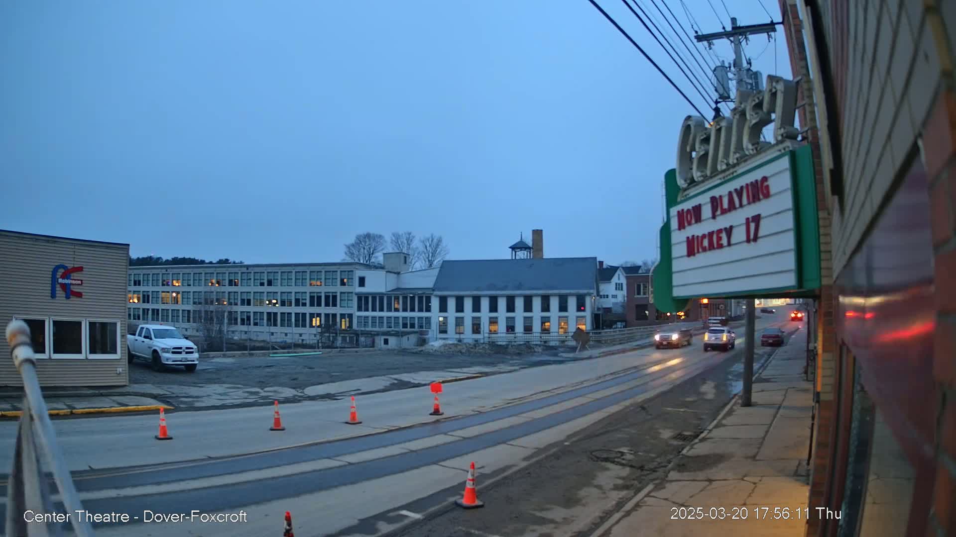 A mostly empty street scene at dusk shows several buildings, including a large multi-story structure, a smaller building with a marquee, and a white pickup truck parked near the curb, with a few traffic cones visible.