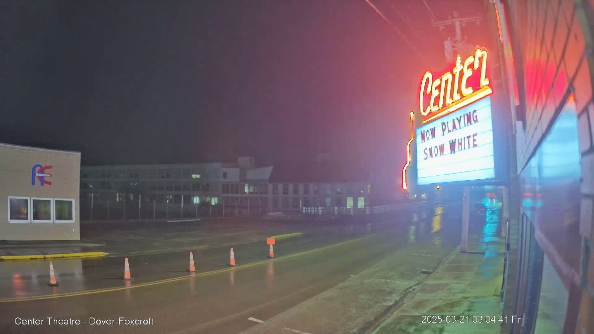 A wet, nighttime street scene shows a movie theater marquee advertising "Snow White," with a building and traffic cones visible in the background.