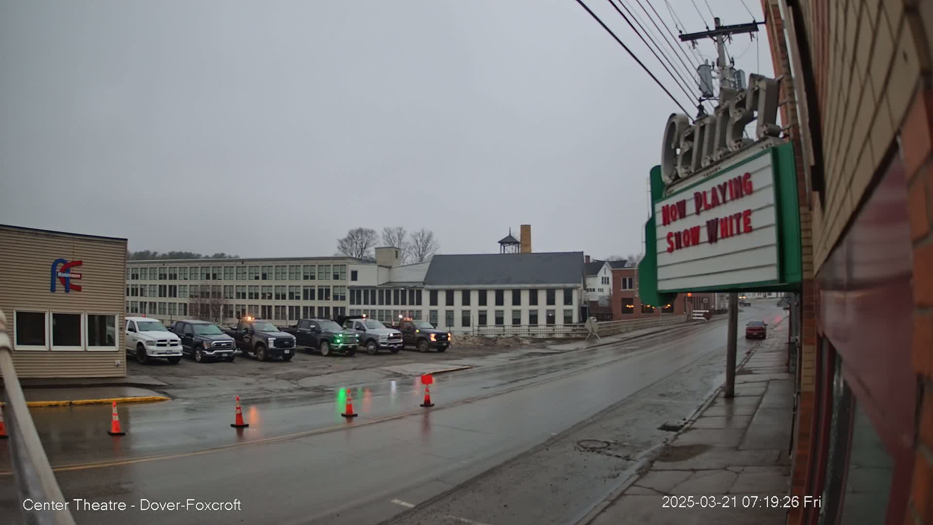 Several pickup trucks are parked in front of buildings on a rain-slicked street under an overcast sky.