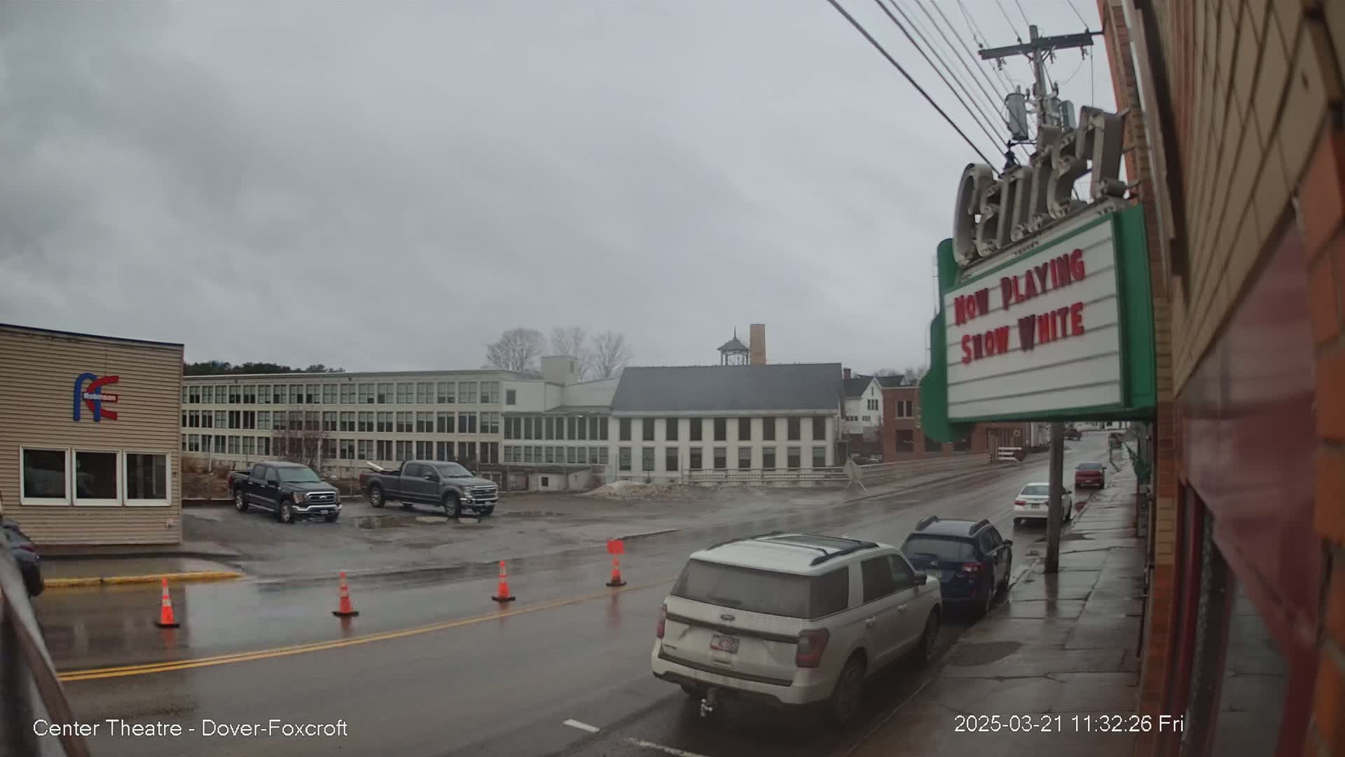 A wet street scene shows several vehicles parked and driving past buildings on an overcast day.