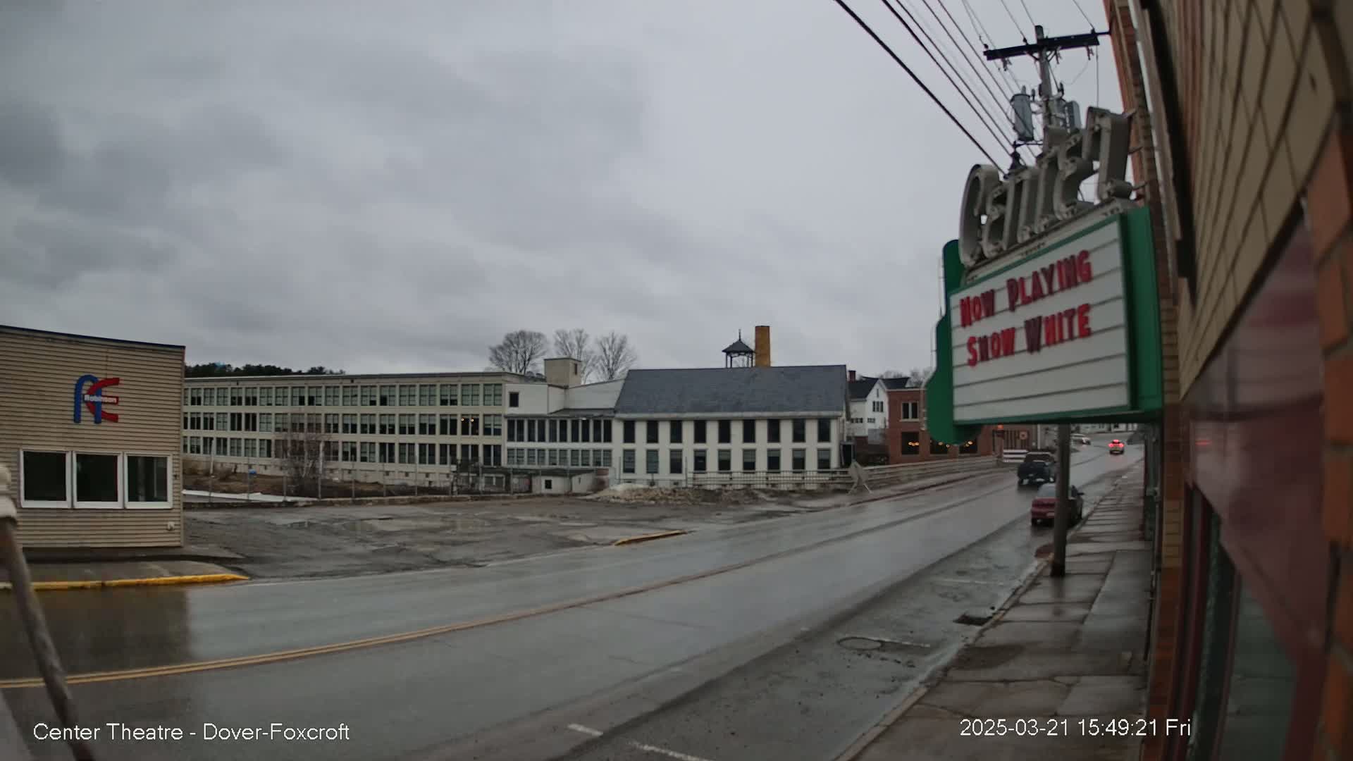 A wet street scene on an overcast day shows a long, multi-story building, a smaller building with a cupola, and a movie theater marquee advertising "Snow White".