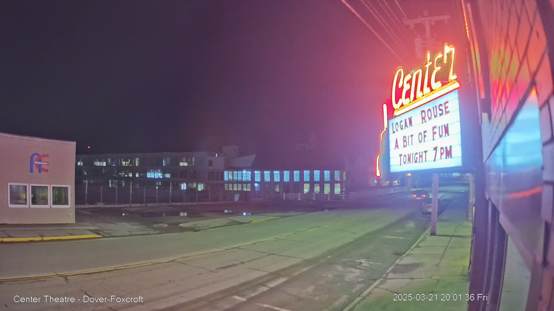 A nighttime street scene shows a theater marquee advertising a show, with a building and a vehicle visible in the distance under a dark sky.