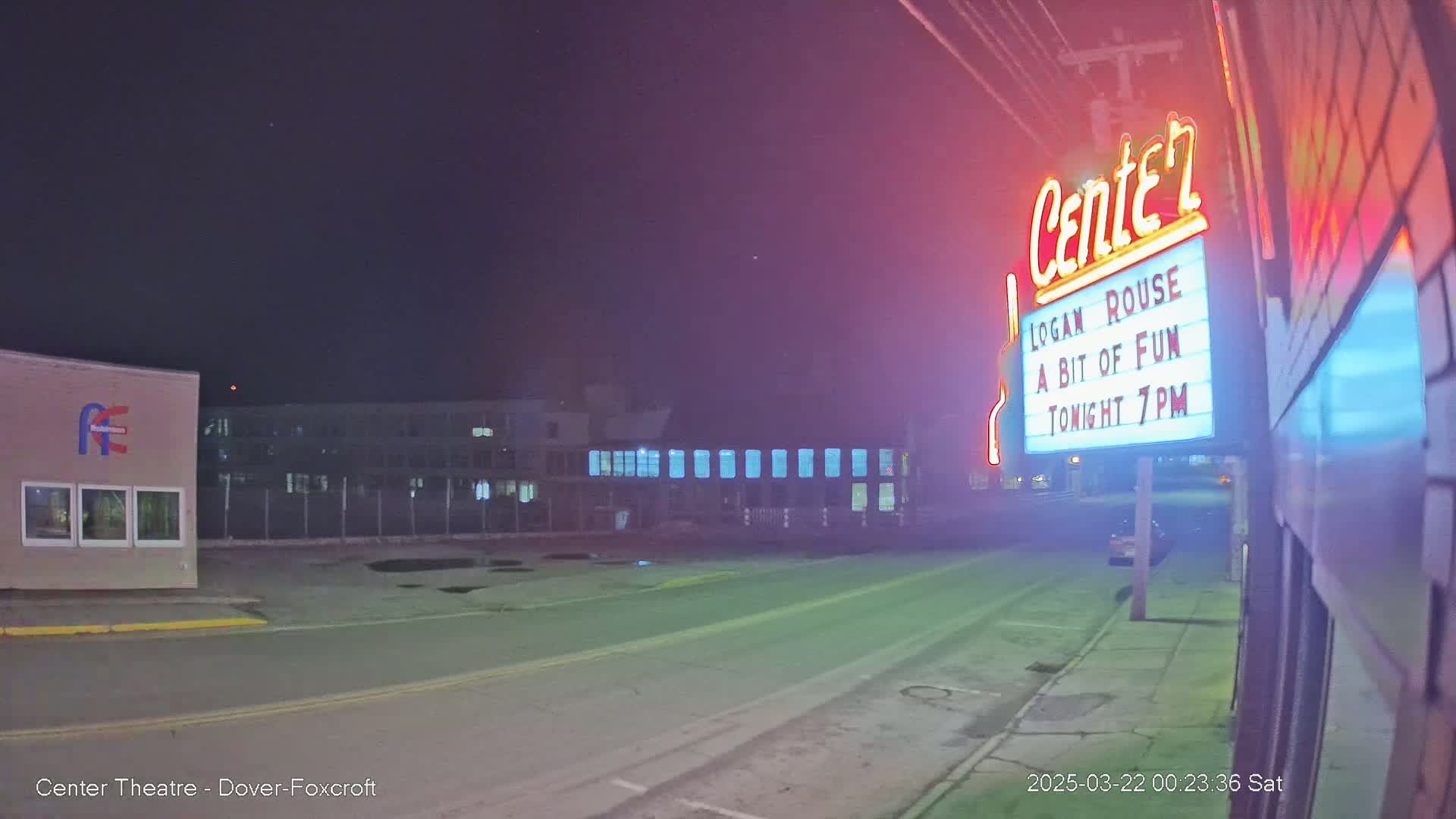 A nighttime street scene shows a theater marquee advertising a show, with a building and empty parking lot visible in the background under a clear, dark sky.