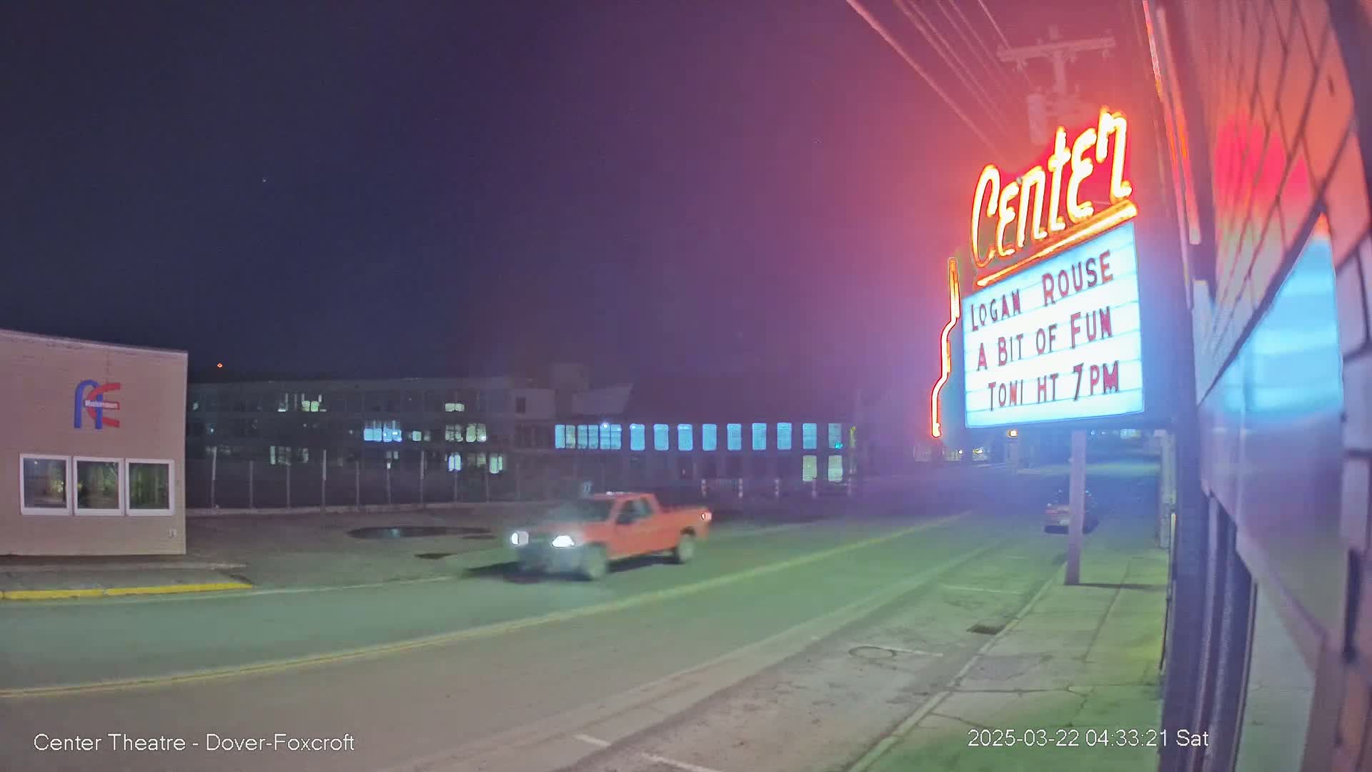 A red pickup truck drives past a movie theater marquee at night under a clear, dark sky.