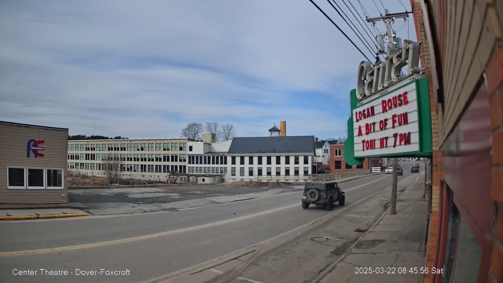 A partly cloudy day reveals a street scene with a dark-colored Jeep driving past older buildings, including a large multi-story factory and a smaller building with a marquee.
