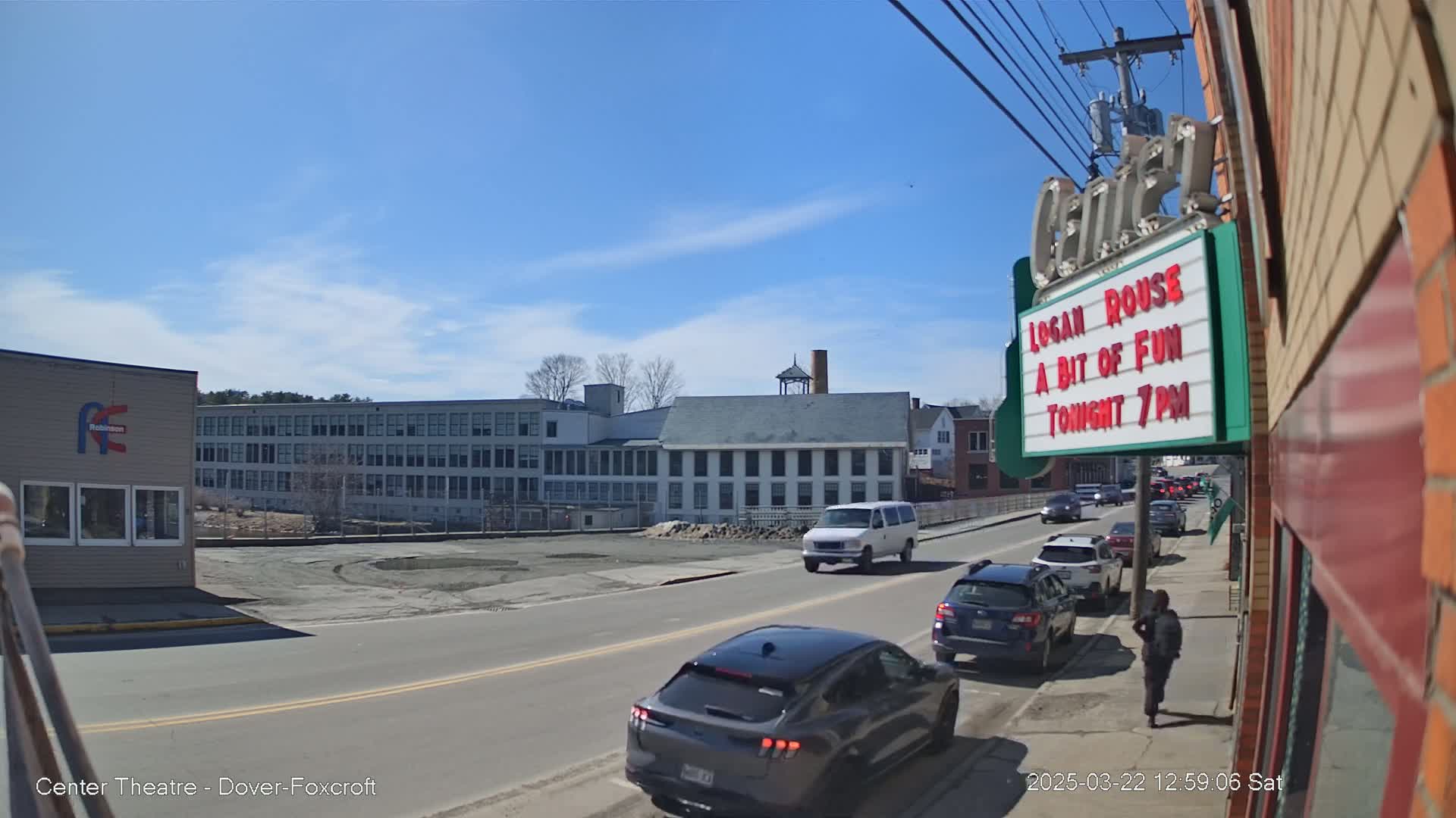 Under a clear blue sky, cars drive down a street past a large white building and a theater marquee advertising a show.