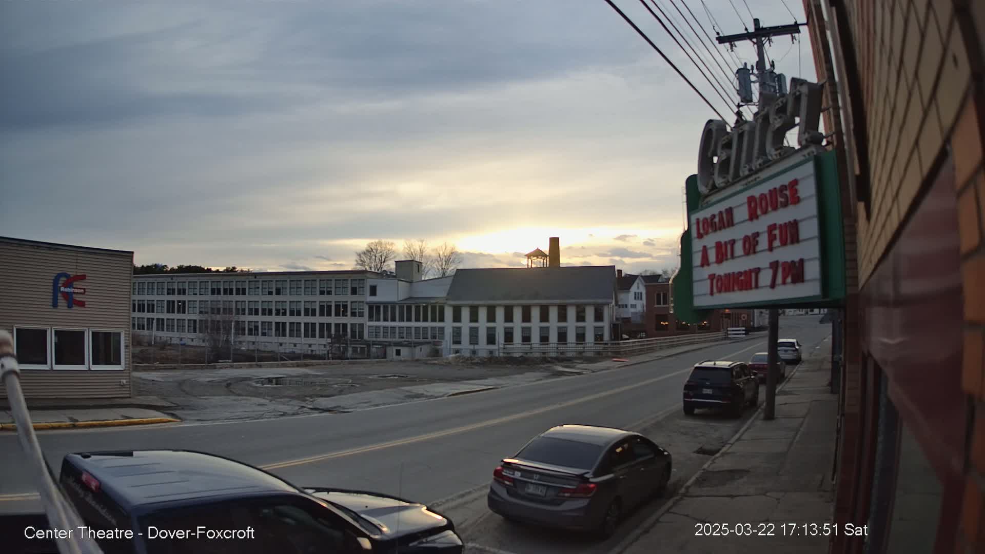 A street scene at sunset shows several cars parked along a road in front of buildings, including a large factory and a theater marquee.