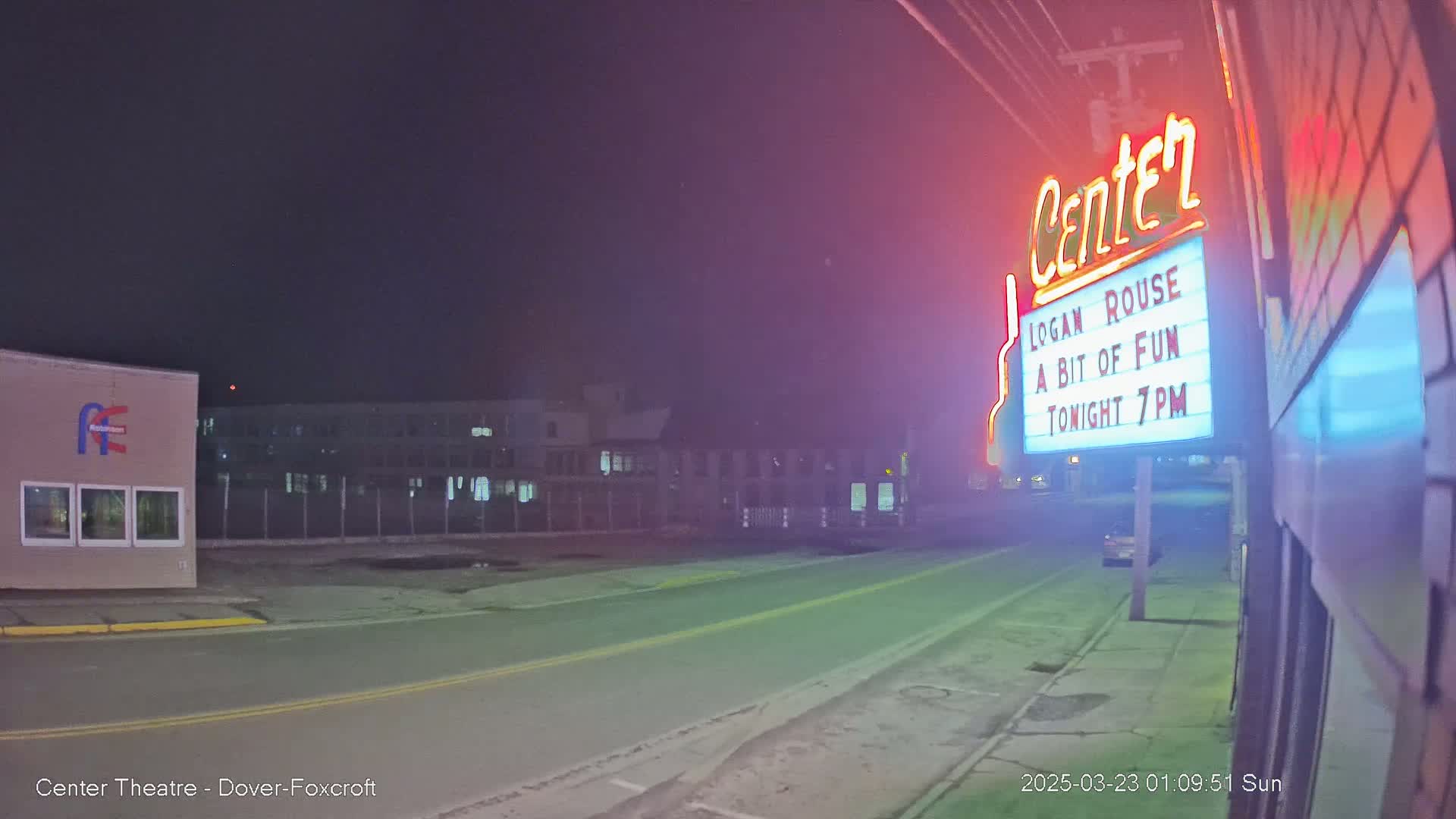 At night, a neon sign illuminates a street scene showing a theater marquee advertising a show, with a mostly empty road and buildings visible in the background.