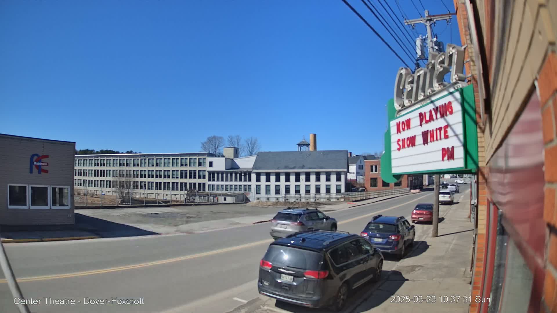 A sunny day reveals a street scene with several cars parked and driving,  passing by industrial buildings and a theater showing "Snow White".