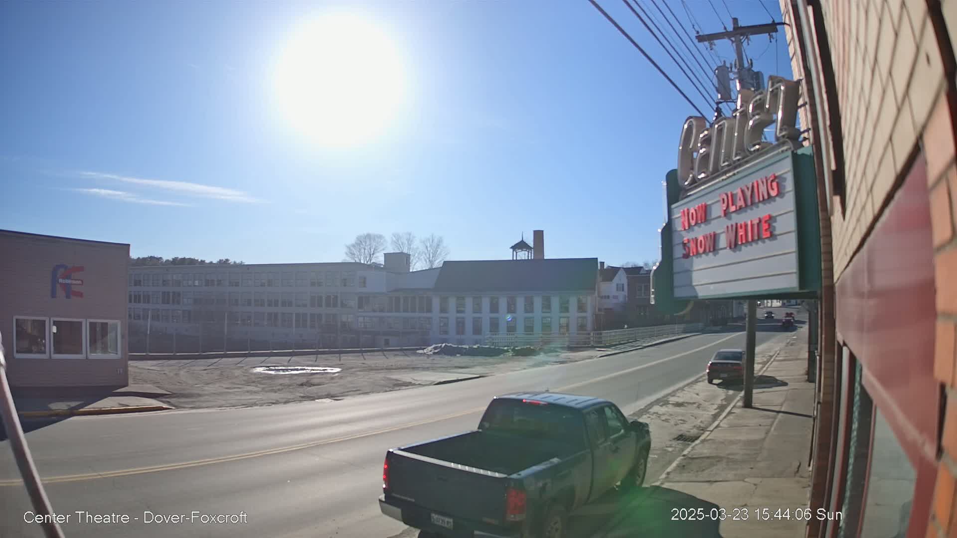 A sunny day reveals a street scene with buildings, including a movie theater marquee, a pickup truck, and other vehicles; a large industrial building is visible in the background.