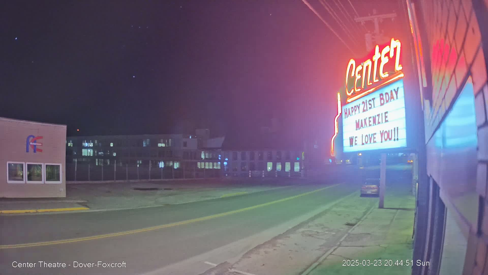 A nighttime street scene shows a building with a marquee displaying a birthday message, a roadway, and other buildings in the background under a clear, starry sky.