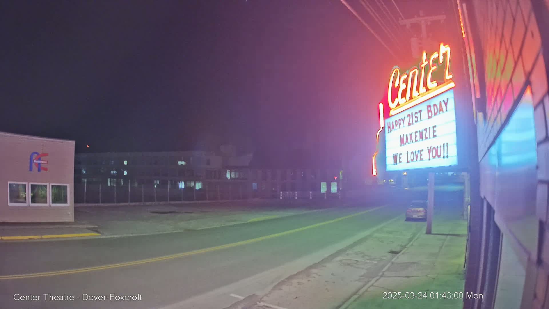 A nighttime street scene shows a building with a lighted sign that says "Happy 21st Birthday Mackenzie We Love You!!"  and a car parked on the road.