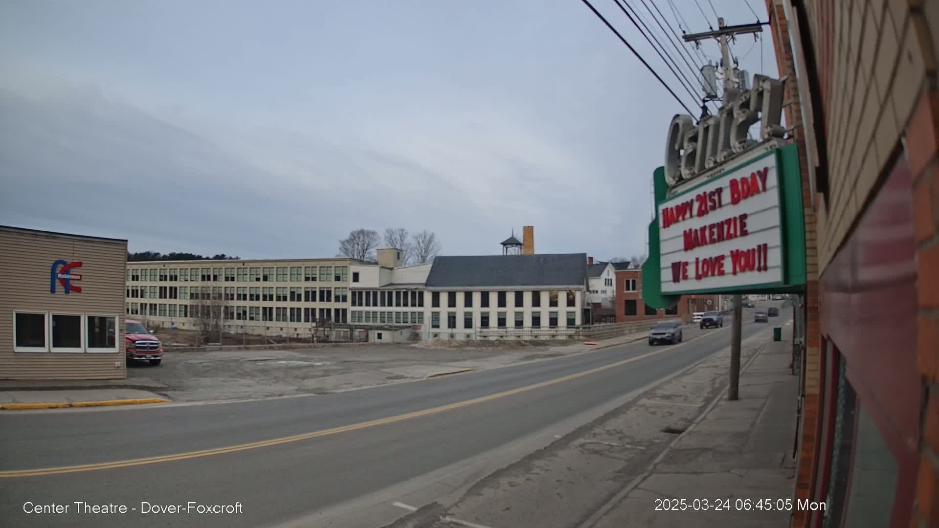 A mostly cloudy day reveals a street scene with several buildings, including a large factory and a smaller building with a marquee, and a few cars are visible on the road.