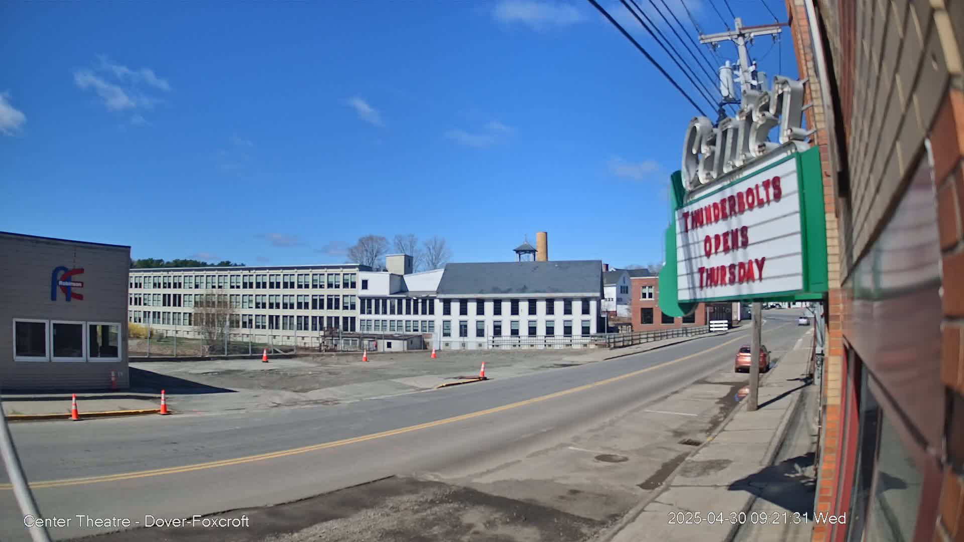 A sunny day reveals a street scene with several industrial buildings, a vacant lot, and a car parked on the side of the road.