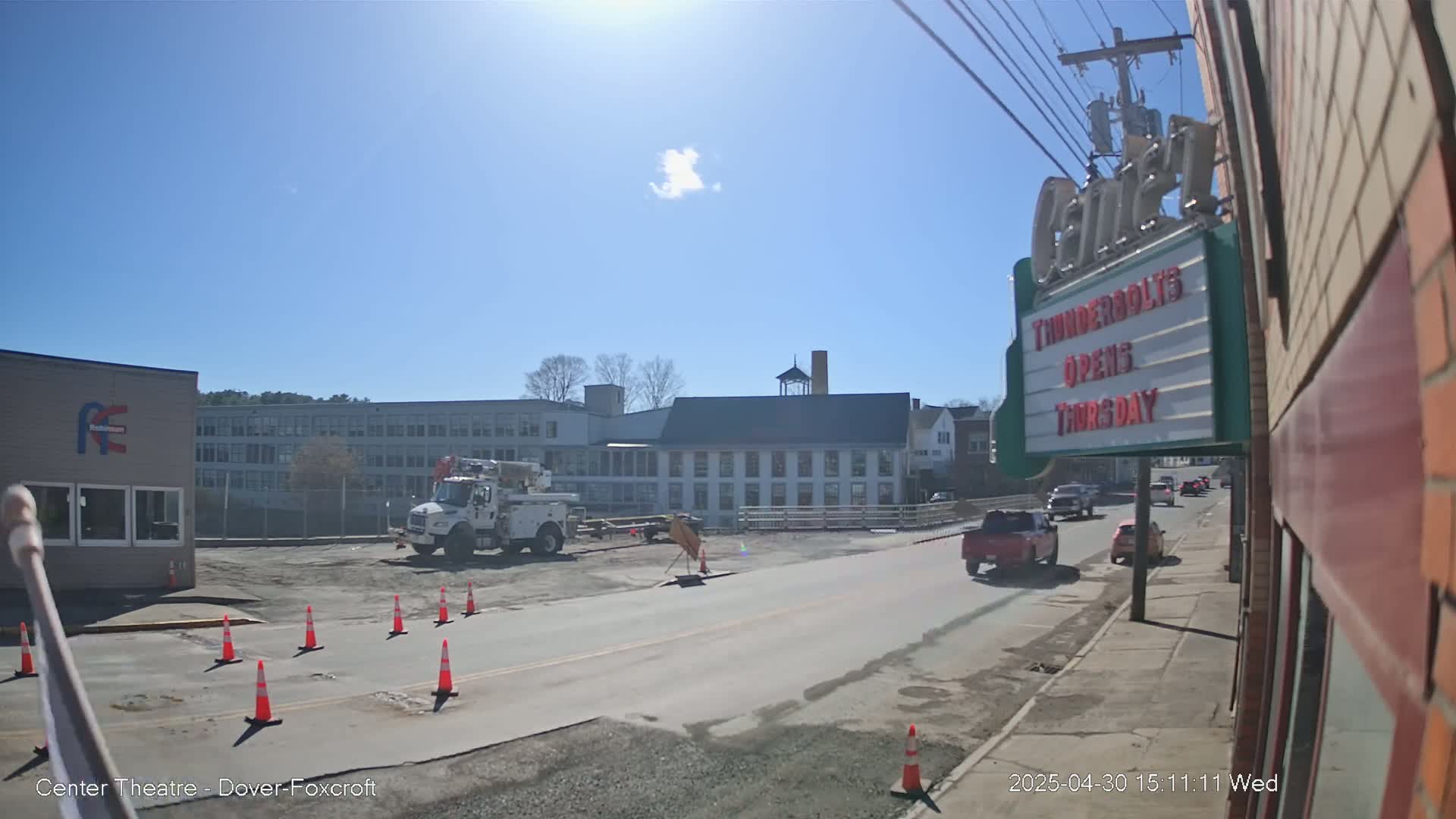 A sunny day reveals a street scene with construction cones and equipment in a cleared area near buildings, including a large multi-story structure and a smaller building with a marquee.