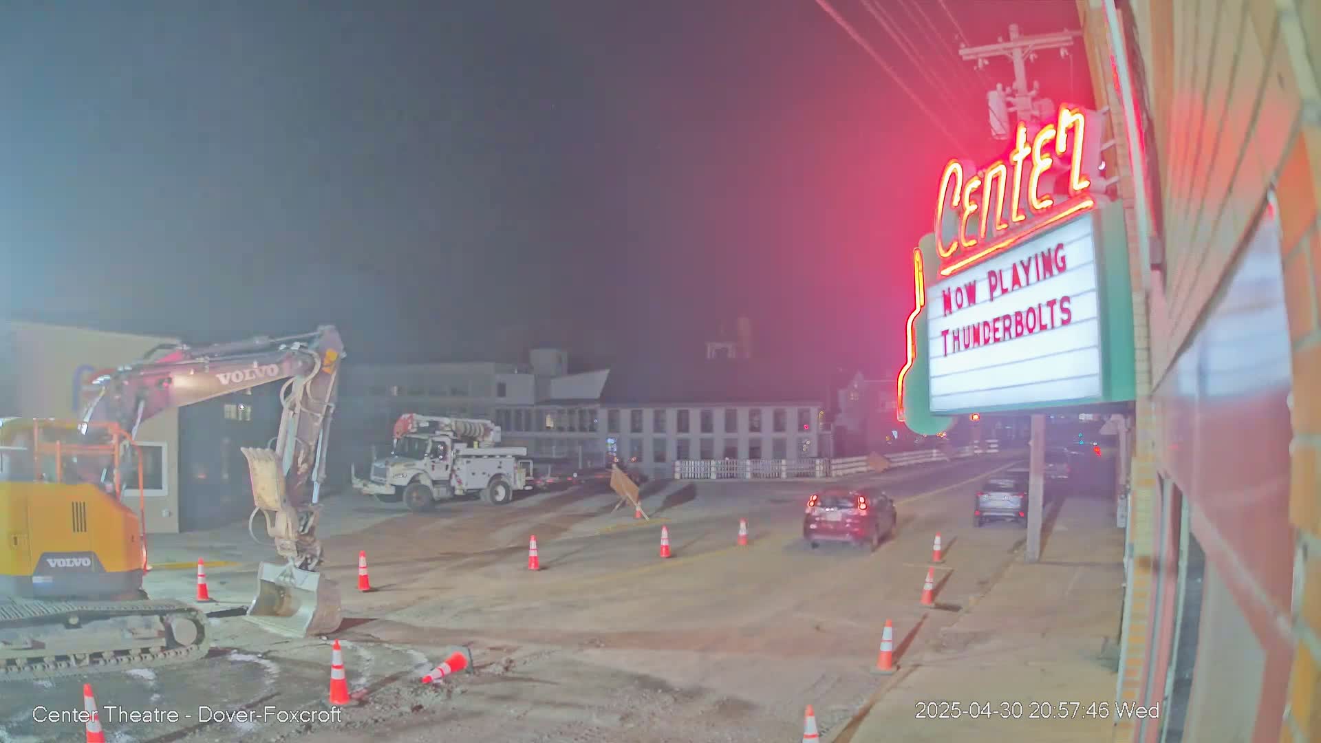 Under a hazy, dark night sky, road construction is underway near a movie theater marquee.