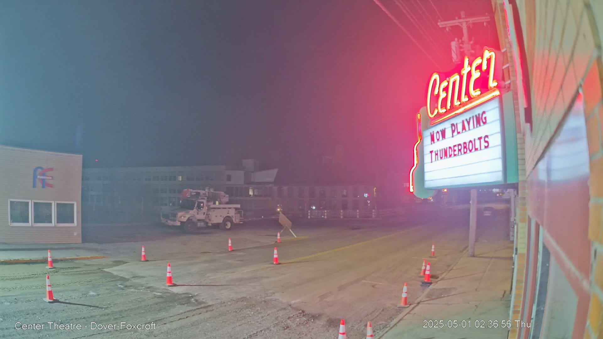 A hazy night scene shows a street with traffic cones, a utility truck, and a movie theater marquee illuminated in red neon.
