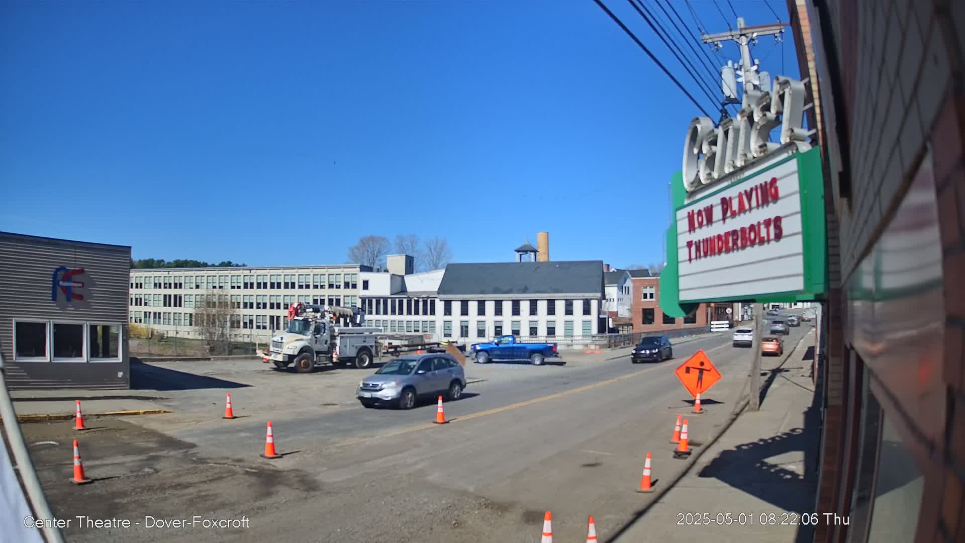 Under a clear blue sky, a street scene shows several vehicles, including a utility truck, a pickup truck, and a silver SUV, with construction cones indicating roadwork in progress near some buildings; one building is a theater.