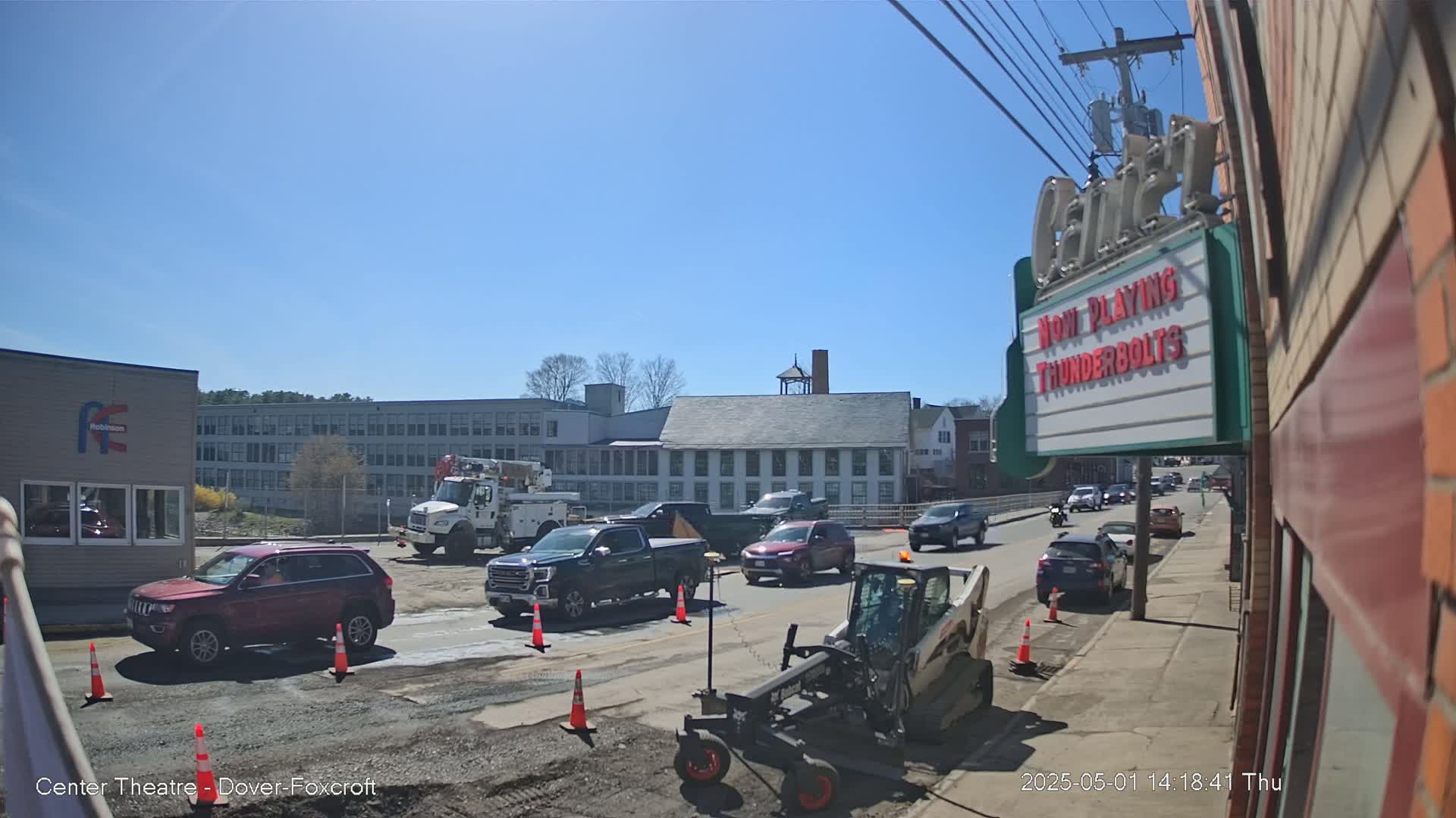 Under a clear blue sky, road construction is underway on a street lined with buildings, including a movie theater, with several vehicles stopped in traffic.