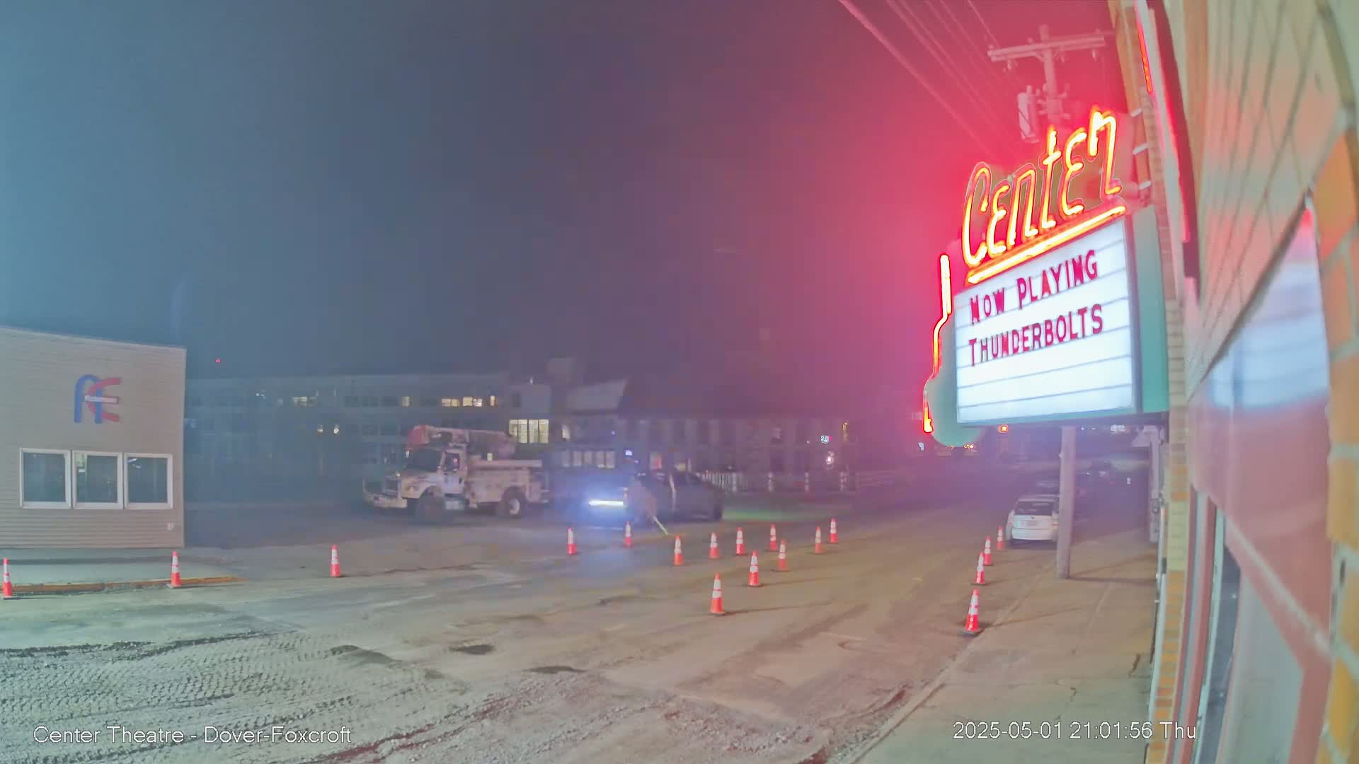 A nighttime, hazy scene shows a street with a few vehicles, including a utility truck, and orange traffic cones in front of a movie theater marquee.