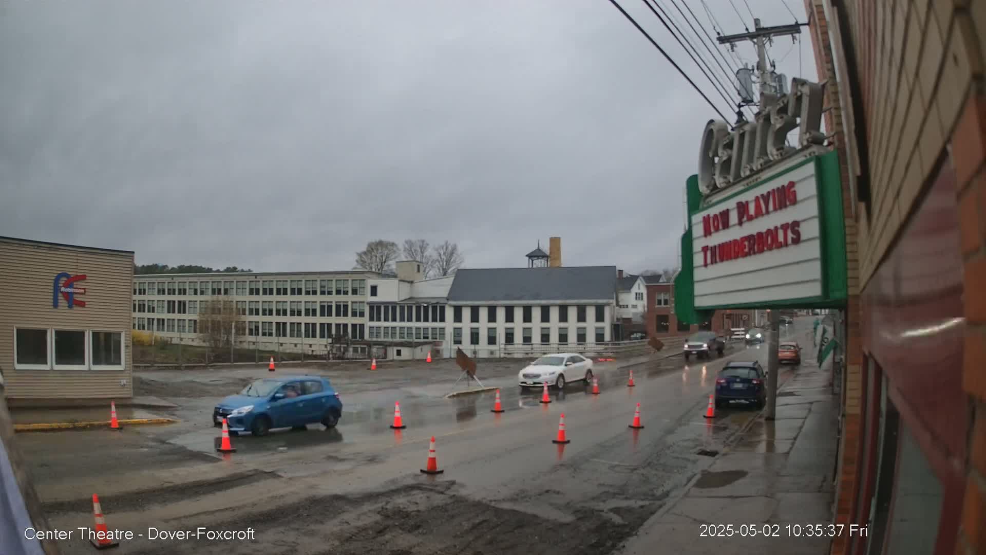 A wet street scene shows several cars driving past orange traffic cones in front of various buildings on an overcast day.