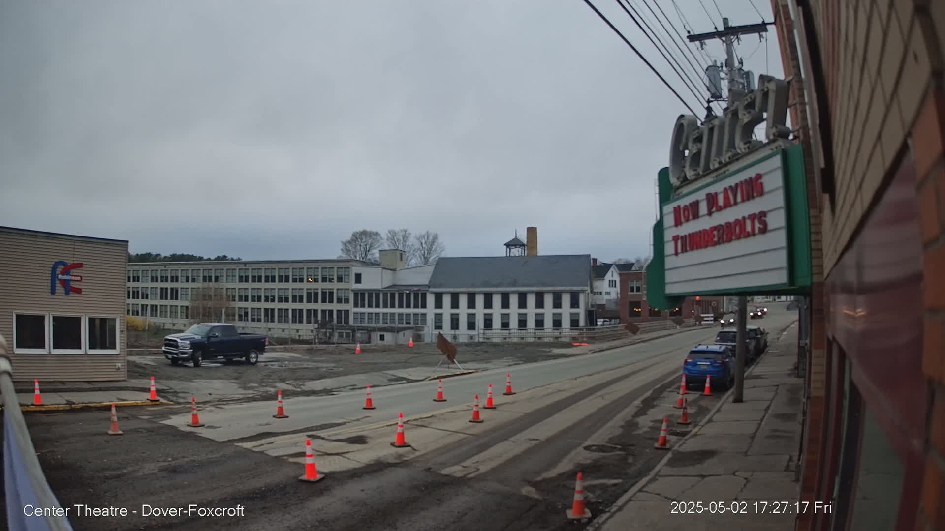 A street scene on an overcast day shows a road under construction with traffic cones, a dark-colored pickup truck, several cars parked along the street, and multi-story buildings in the background.